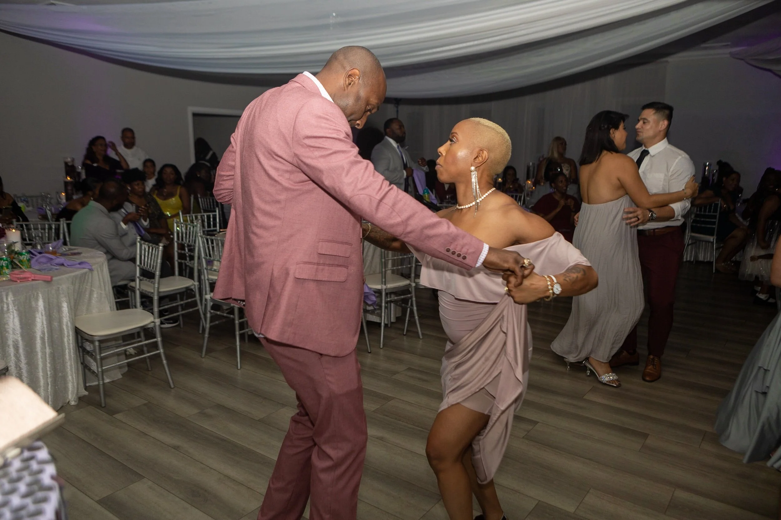 A man and woman dance closely at a wedding reception, with the man in a pink suit and the woman in an off-the-shoulder beige dress, surrounded by other couples and guests in a decorated event hall.