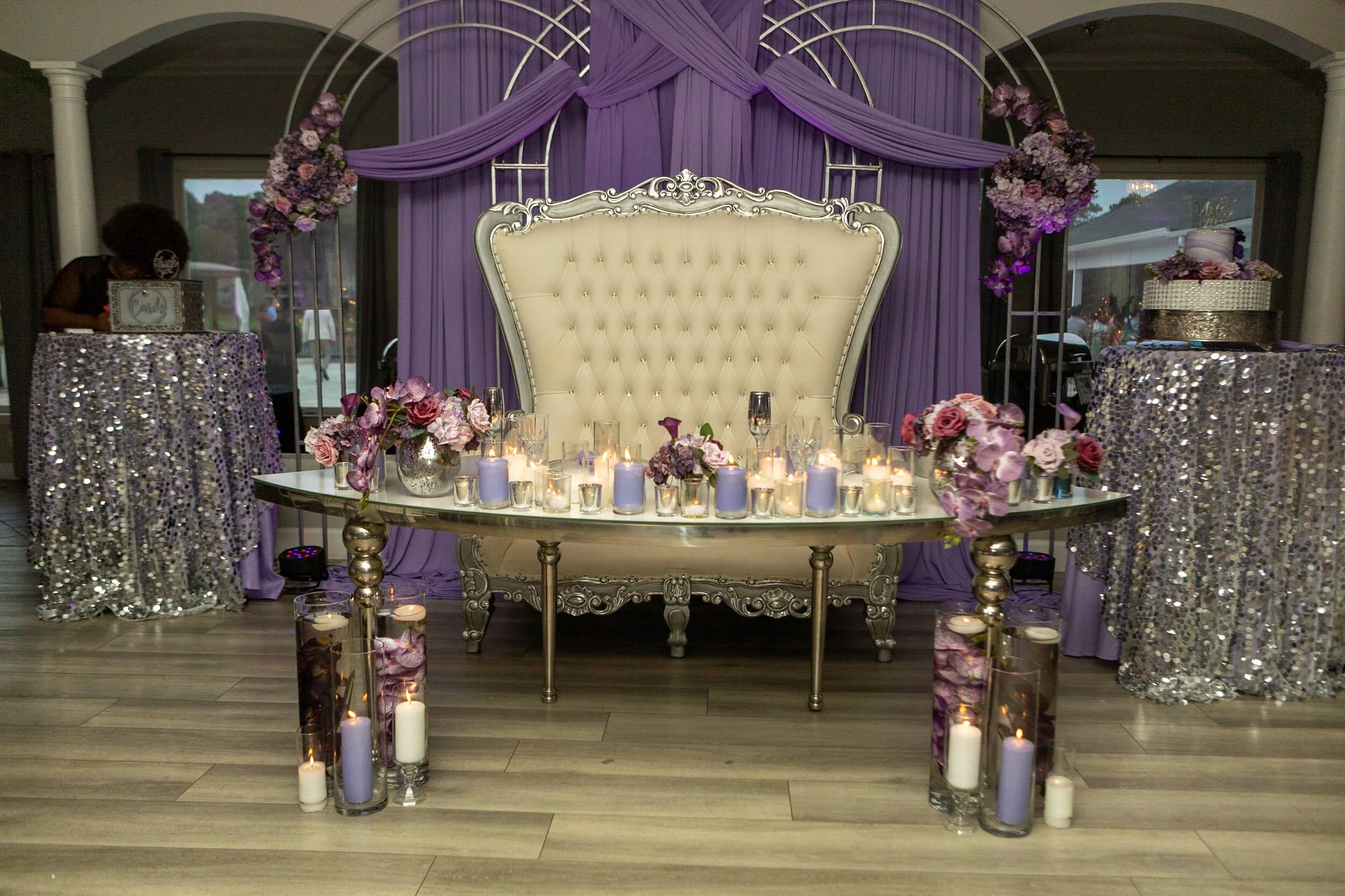 Decorative setup with purple, silver, and white wedding-themed decorations, including a high-backed ornate chair, floral arrangements, candles, and a sequined tablecloth in a reception hall.