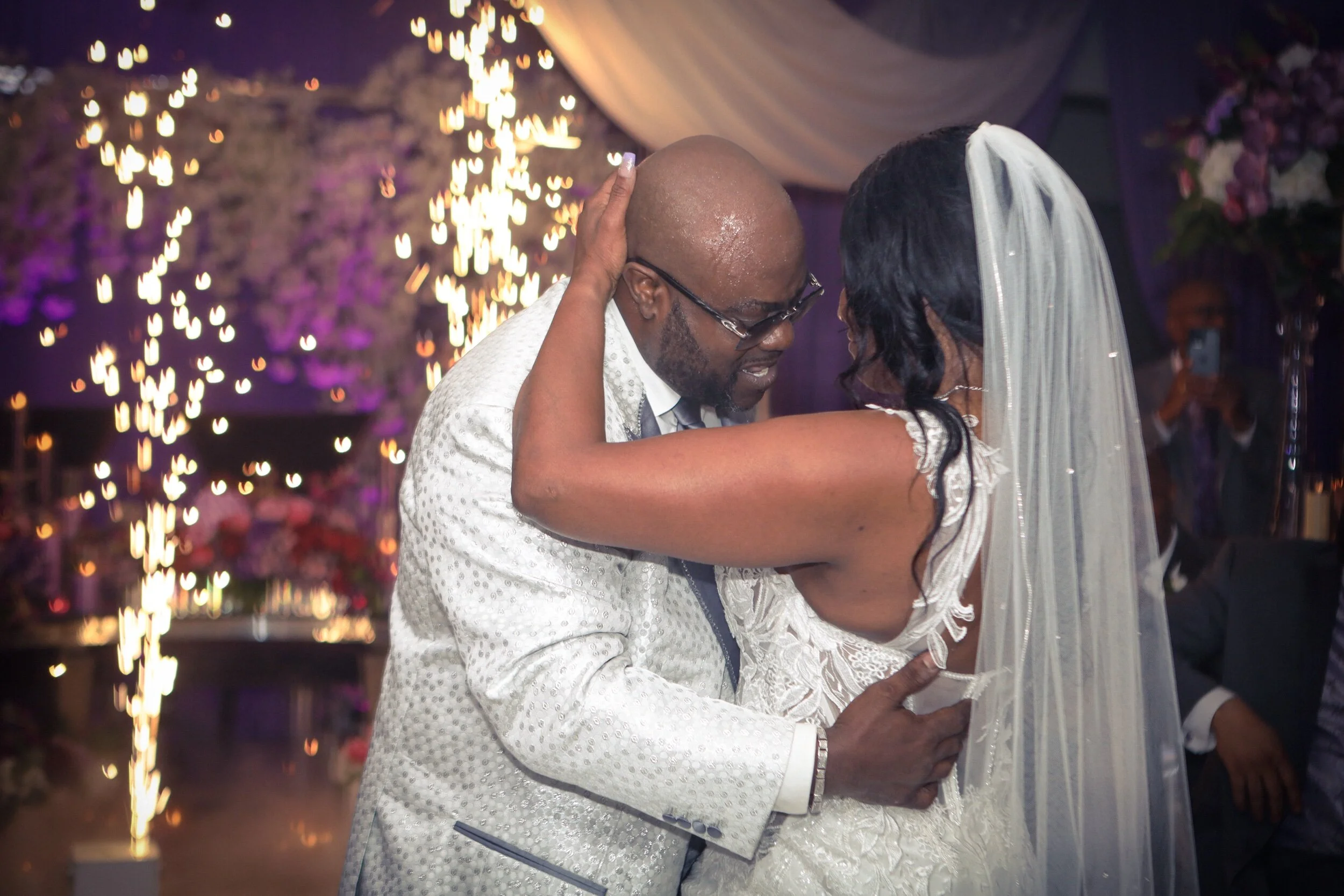 A bride and groom are dancing closely at their wedding reception, with fireworks in the background and purple lighting.