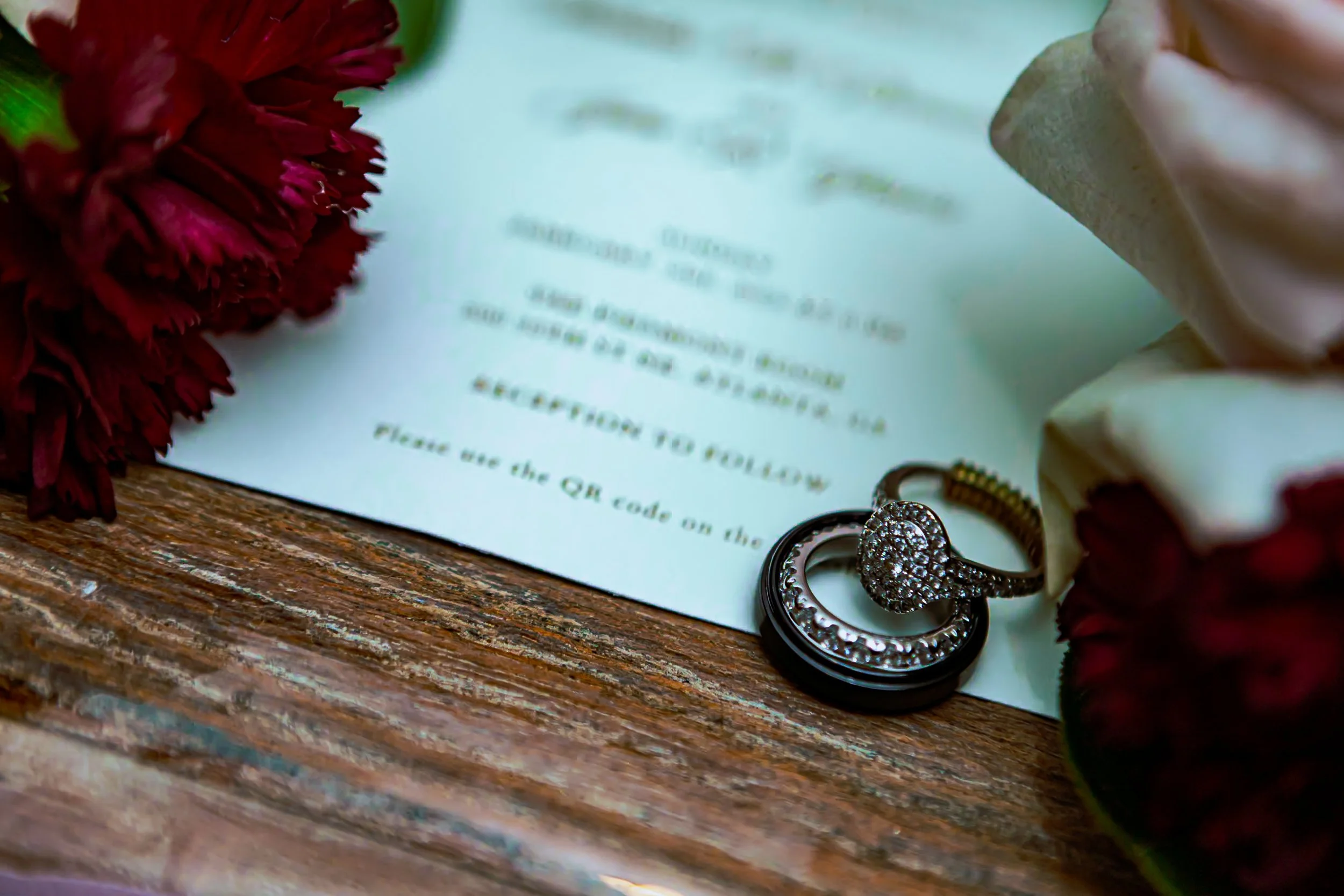 A wedding invitation, a gold wedding band with diamonds, a black ring, a maroon flower, and white roses on a wooden surface.