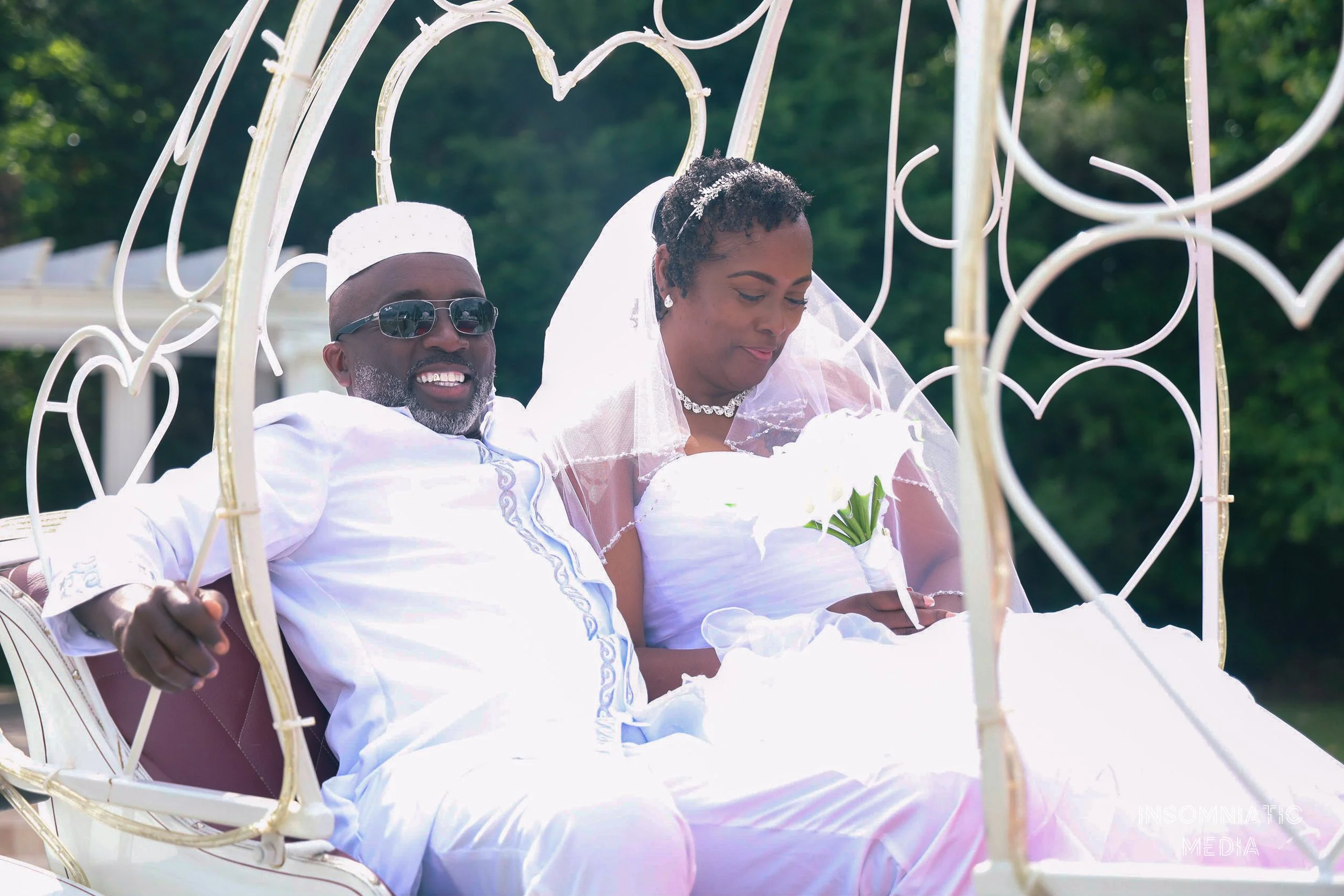 A bride and groom sitting inside a decorative white carriage during their wedding, with the groom wearing sunglasses and a white cap, and the bride holding a white bouquet, outdoors with green trees in the background.
