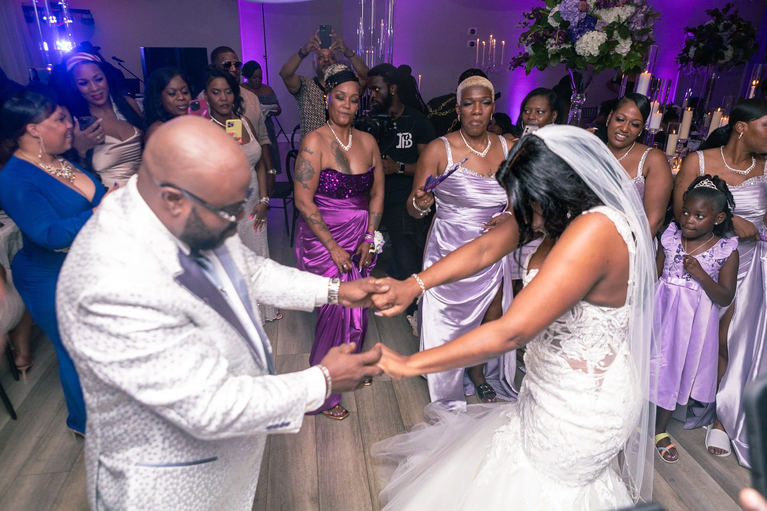 A bride and an older man dance together at a wedding reception surrounded by guests in elegant dresses. The bride wears a white wedding gown with a veil, and the man is dressed in a white jacket. The guests are watching and capturing the moment, with