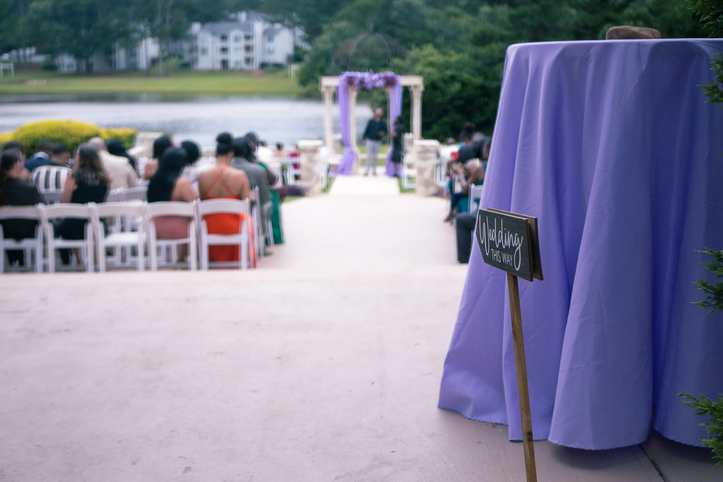 Outdoor wedding ceremony setup by a lake with chairs arranged facing the altar, purple drapery on the side table, and a sign indicating the direction to the wedding.
