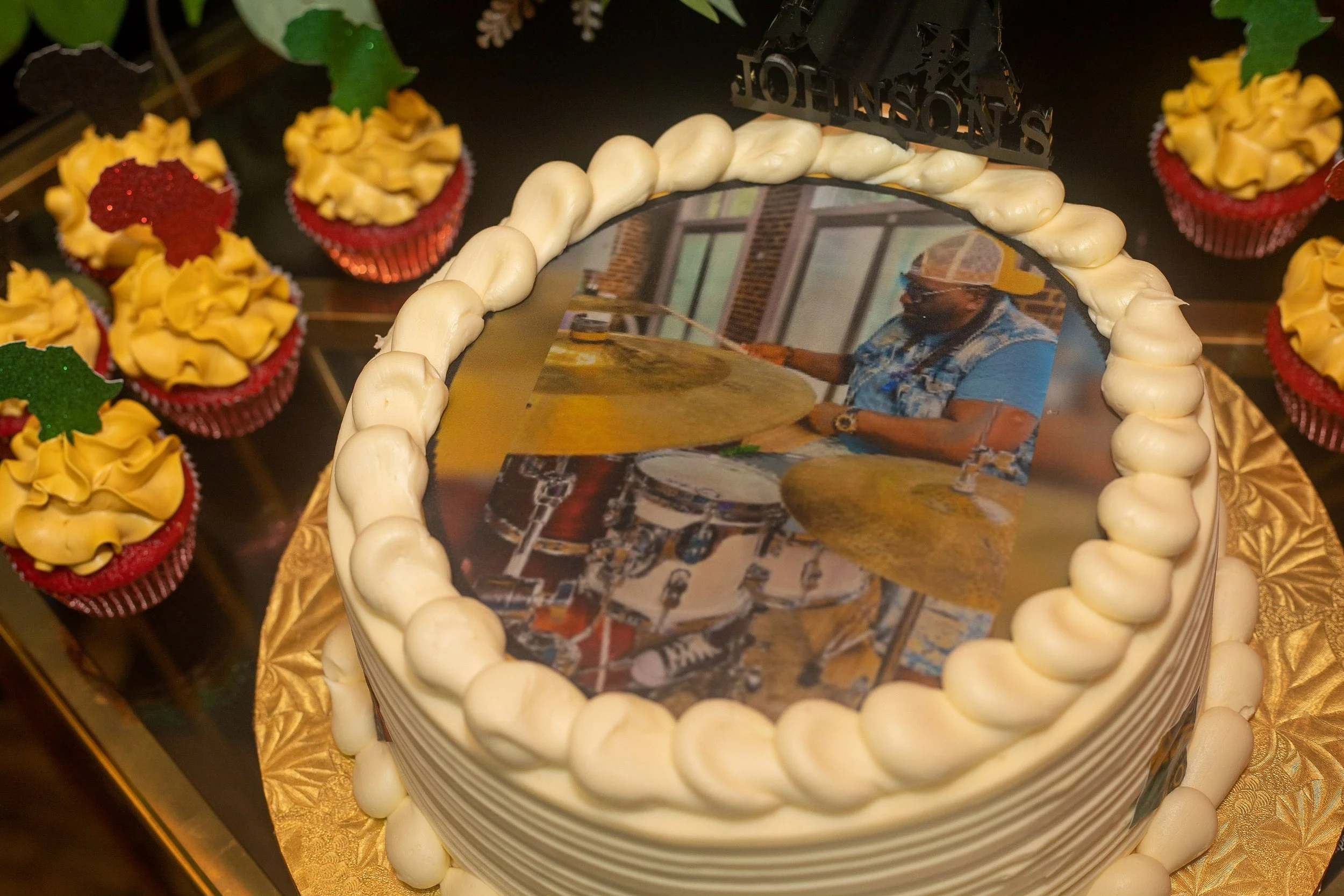 Decorative round cake with an edible photo on top of a drummer playing drums, surrounded by a border of white cream piped in swirls. Several red cupcakes with yellow frosting and holly-shaped green and red toppers are arranged around the cake.