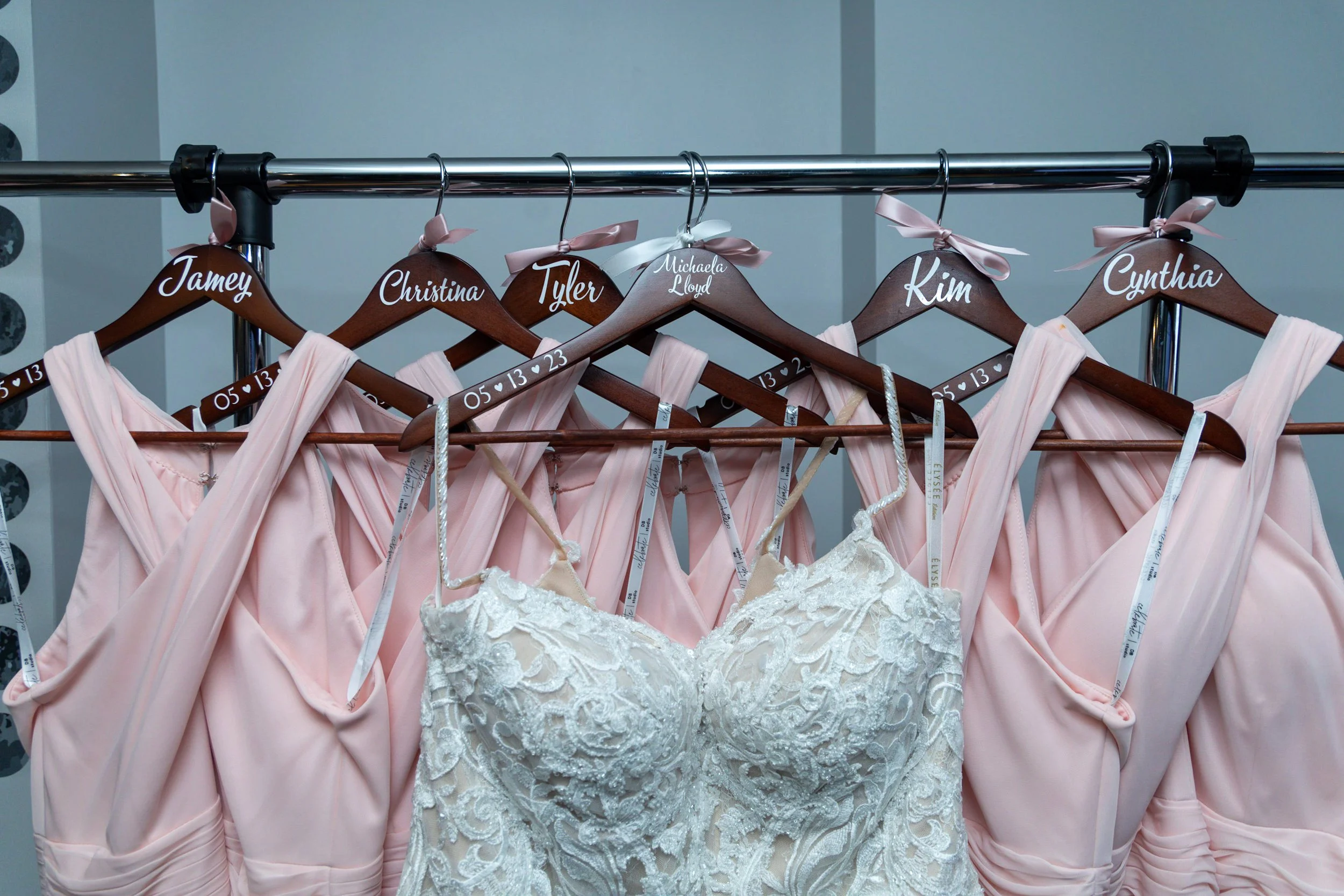 A row of pink bridesmaid dresses and a white lace wedding dress on hangers, hanging on a metal rod. The hangers are wooden with handwritten names on top, such as Jamey, Christina, Tyler, Michaela Lloyd, Kim, and Cynthia. Some hangers have ribbons tie