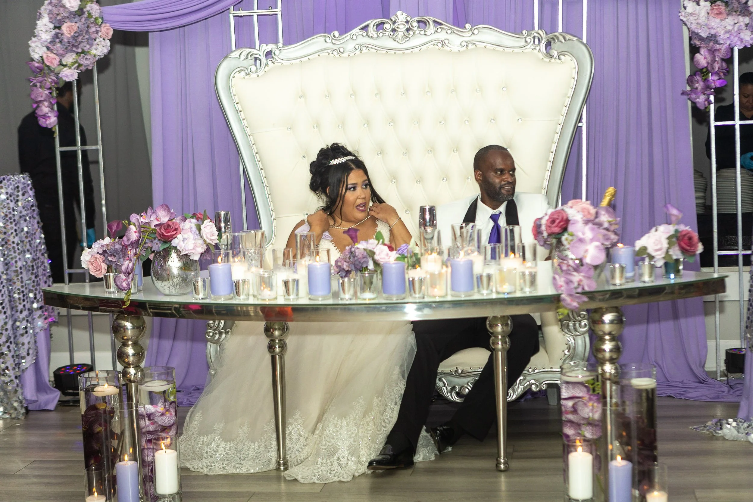 Bride and groom sitting on a fancy white and silver throne-like chair at their wedding reception table decorated with purple and pink flowers and lit candles, with purple drapes in the background.