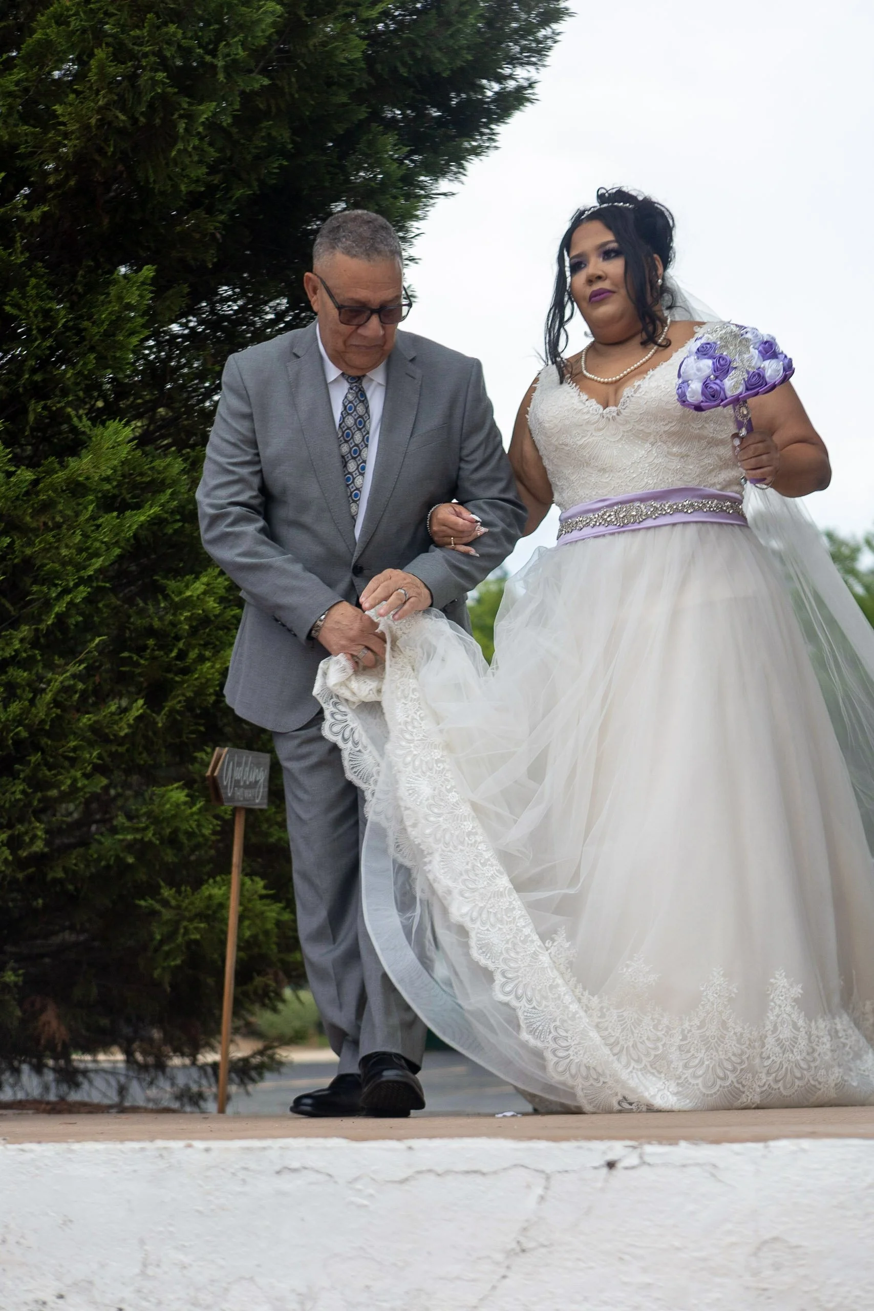 Bride in a white wedding dress and purple sash holding a purple flower, walking with man in a gray suit on an outdoor stage, with greenery in the background.