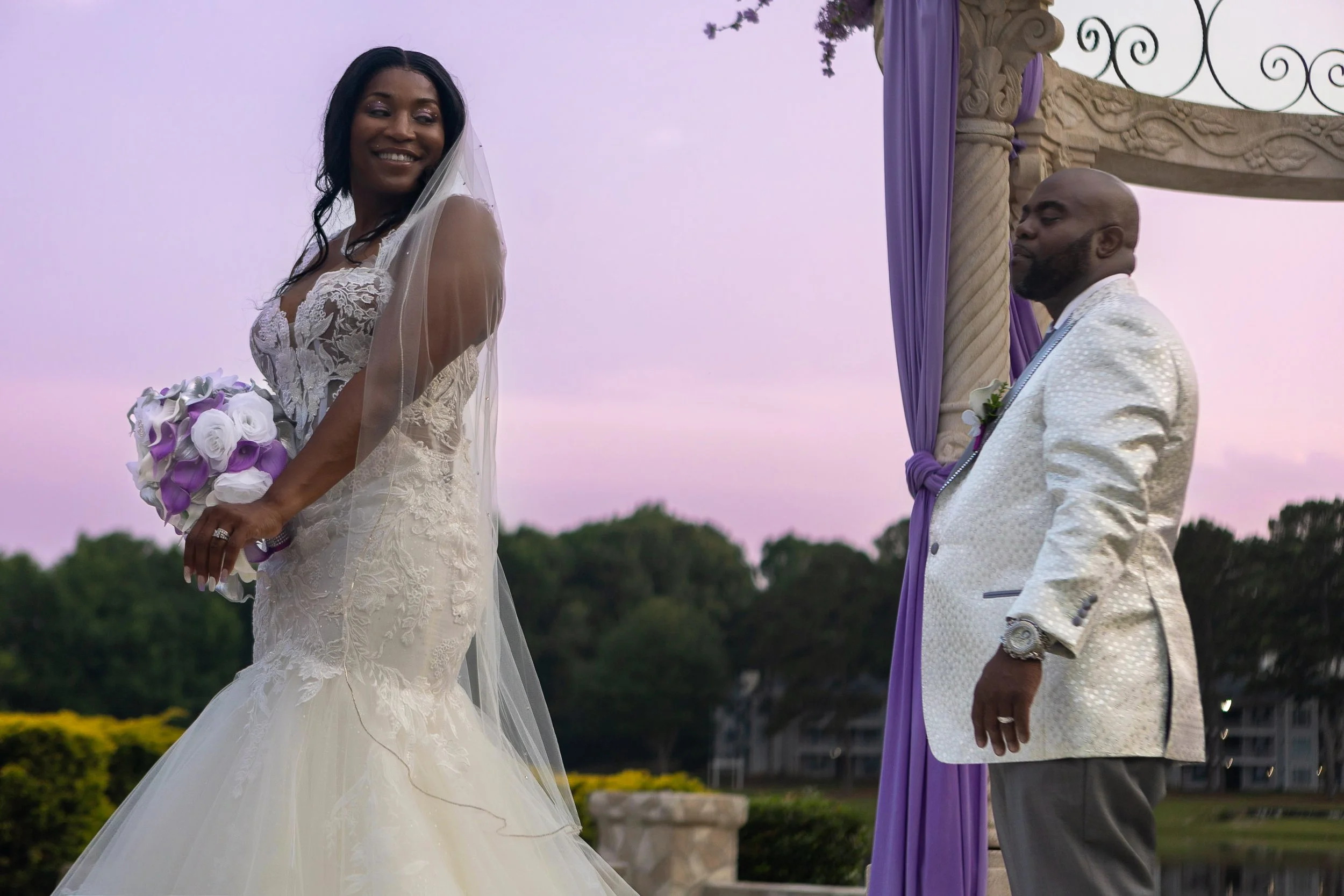 A bride in a white wedding gown holding a bouquet of white and purple flowers, smiling, during her outdoor wedding ceremony at sunset. The groom, dressed in a white suit with a purple sash, stands under a decorated arch with purple drapes.