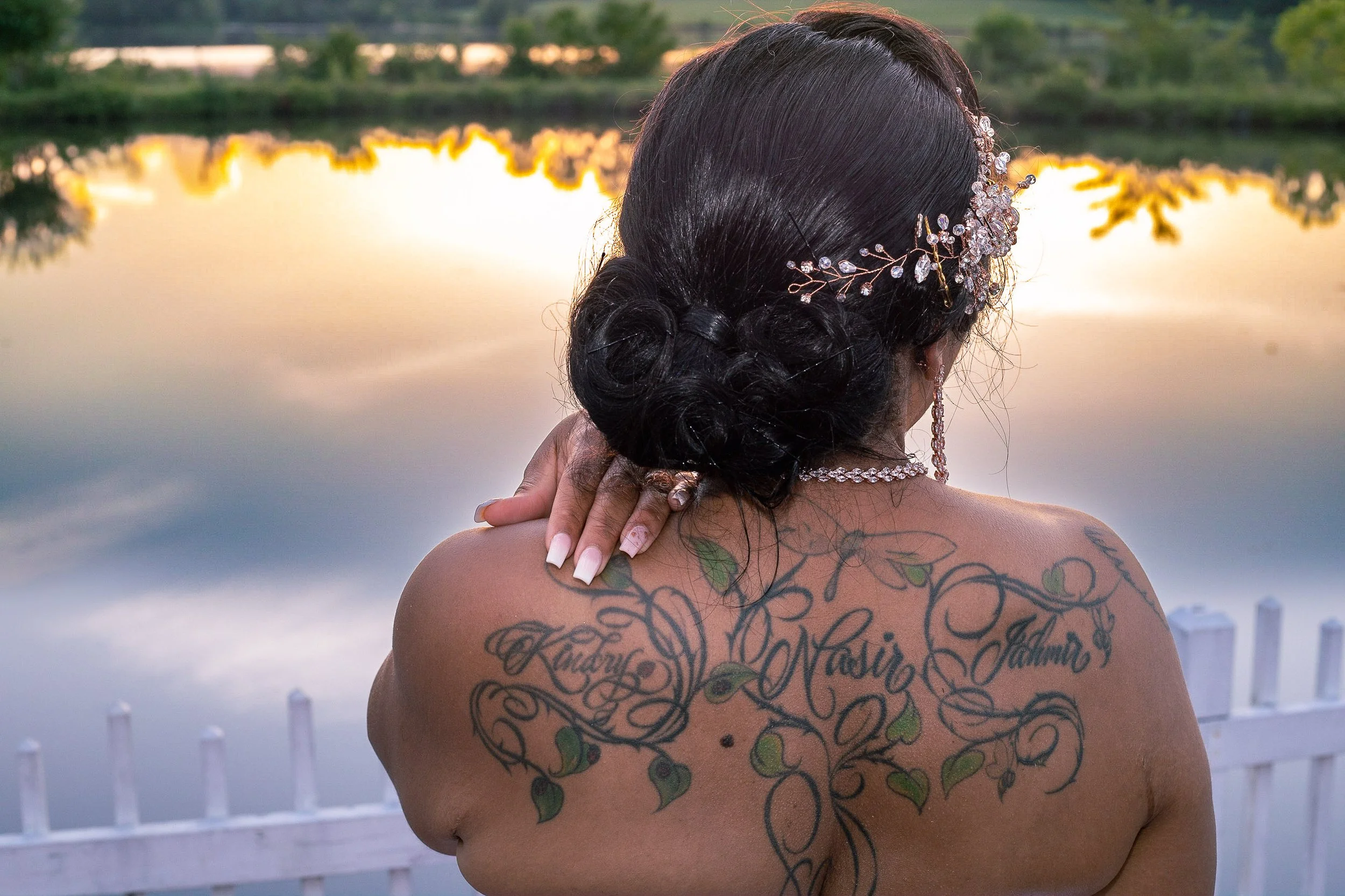 Woman with dark hair styled in an updo, wearing a jeweled headpiece, posing with her back to the camera, overlooking a body of water at sunset. She has tattoos on her back with words and a vine design, and is dressed in jewelry.