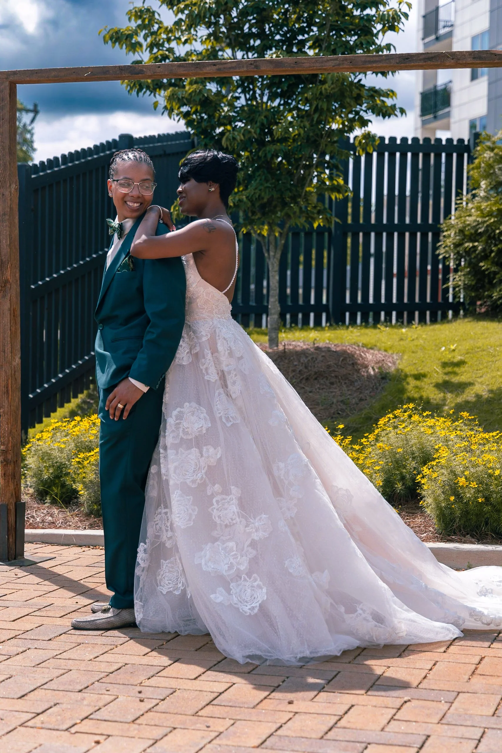 Two women, one in a wedding dress and the other in a suit, embrace outdoors on a paved patio, smiling with a garden and black fence in background.
