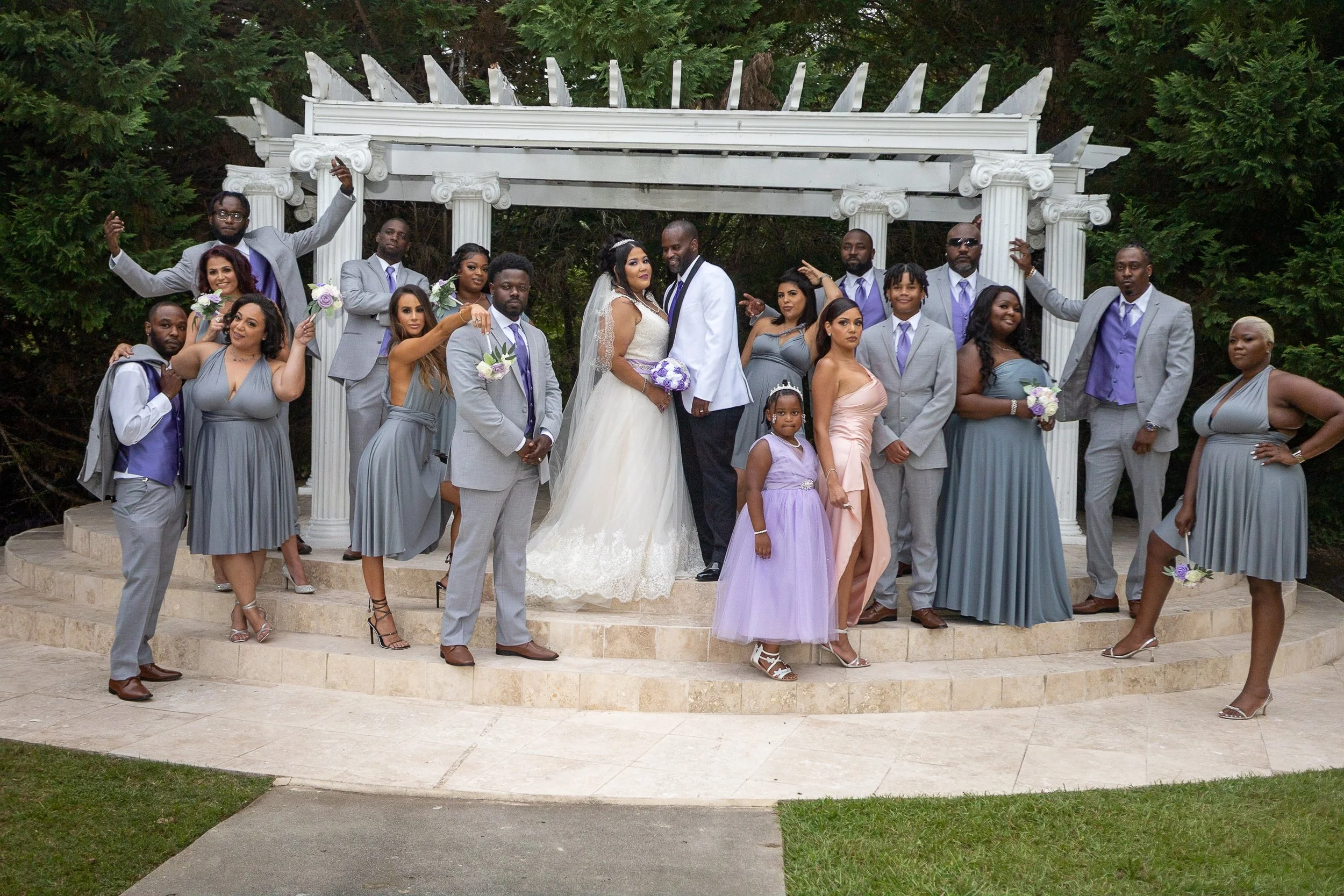 A wedding party with the bride and groom in the center, surrounded by bridesmaids and groomsmen, standing on a circular stone platform with a white classical-style arch. The group is dressed in formal attire, with the women in dresses and the men in 