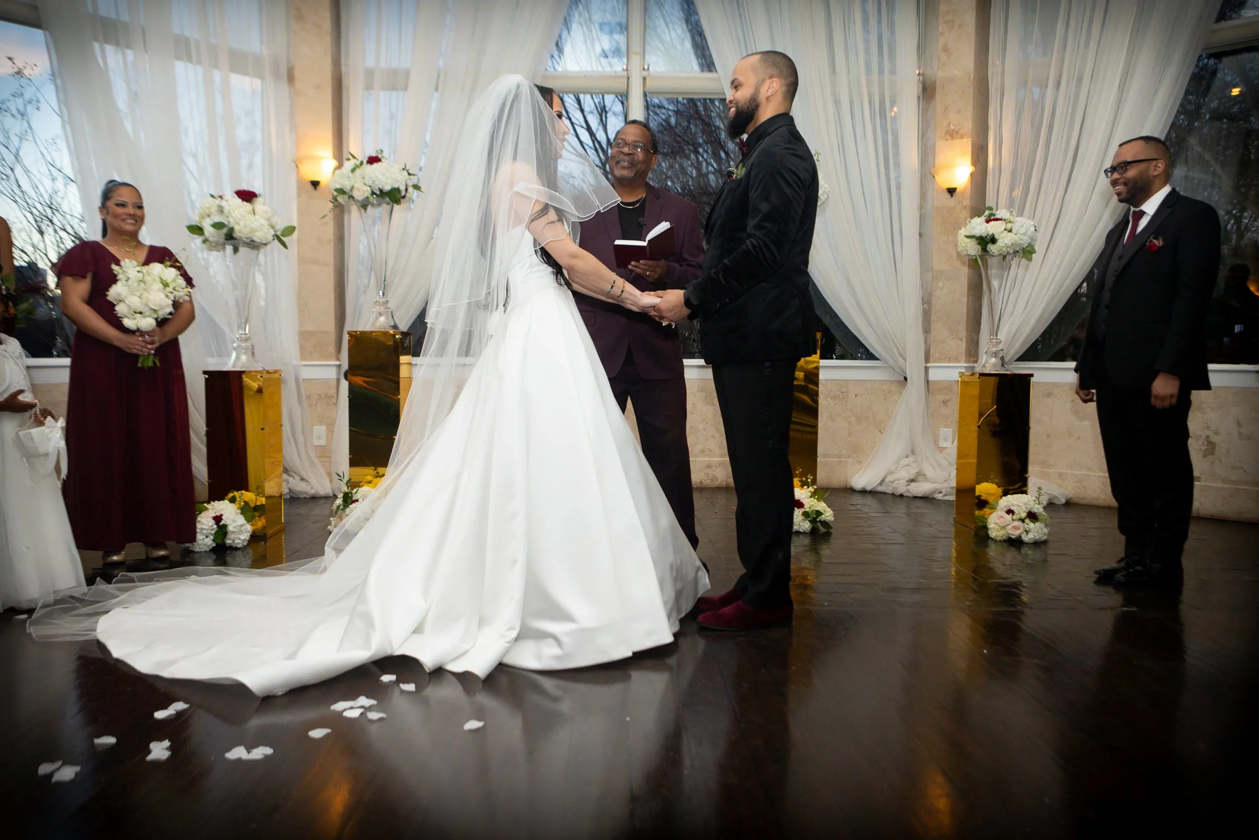 A wedding ceremony with a bride and groom holding hands and facing each other, with an officiant in the middle, surrounded by bridesmaids and groomsmen.