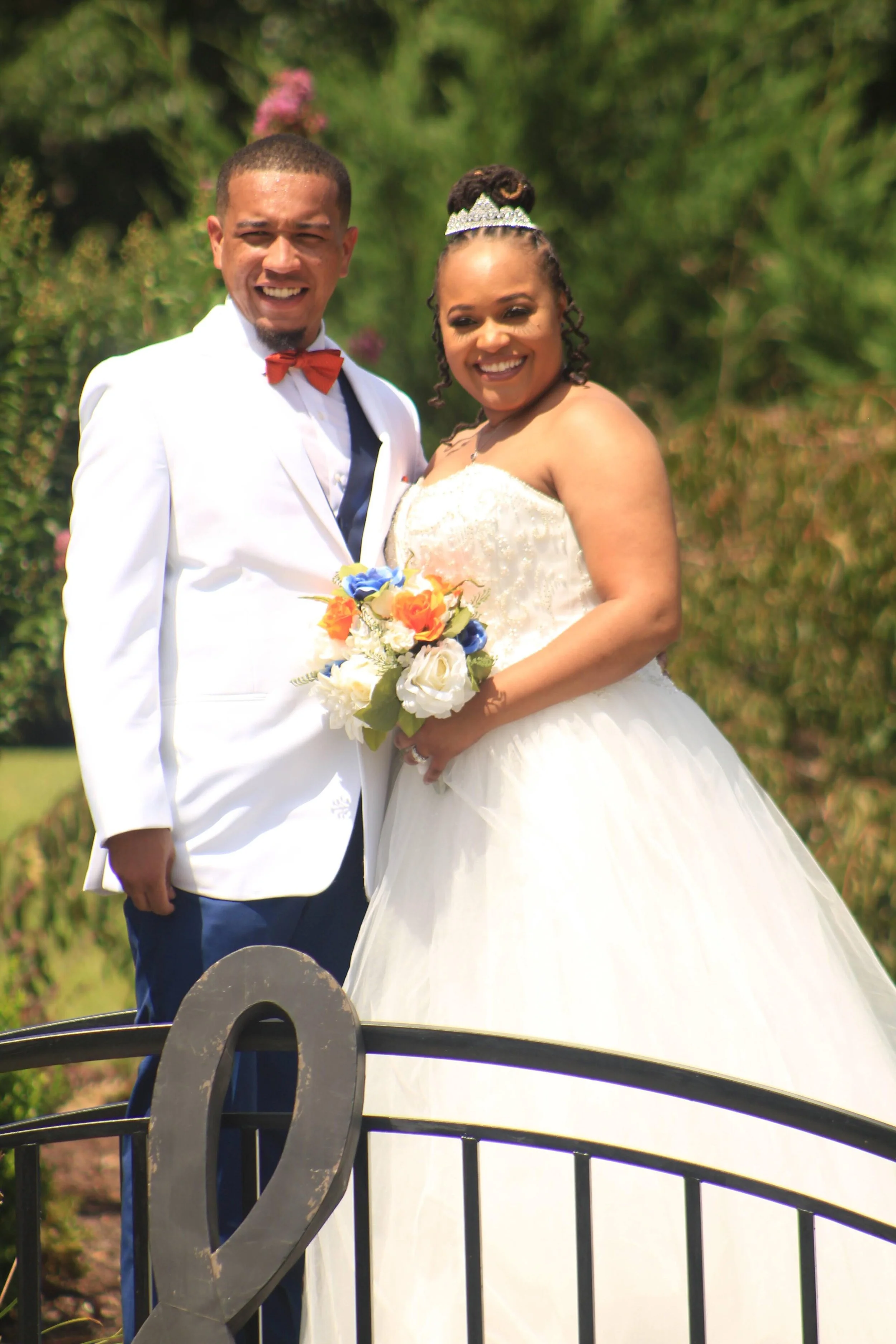A bride and groom on their wedding day smiling outdoors, with the bride wearing a white wedding gown and a tiara, holding a bouquet of colorful flowers, and the groom in a white suit with a red bow tie, standing on a decorative bridge with a heart-sh