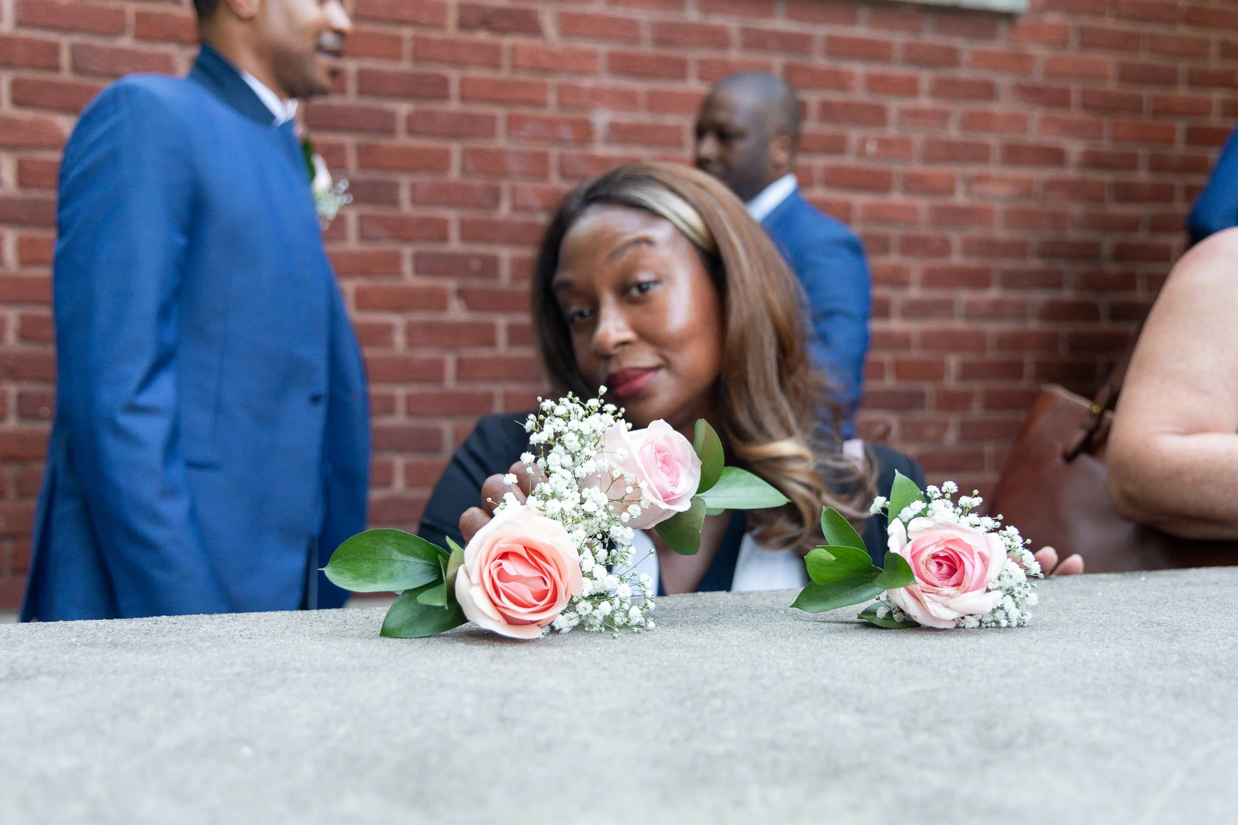 A woman resting her head on a table with pink roses and greenery, with a brick wall and people in formal attire in the background.