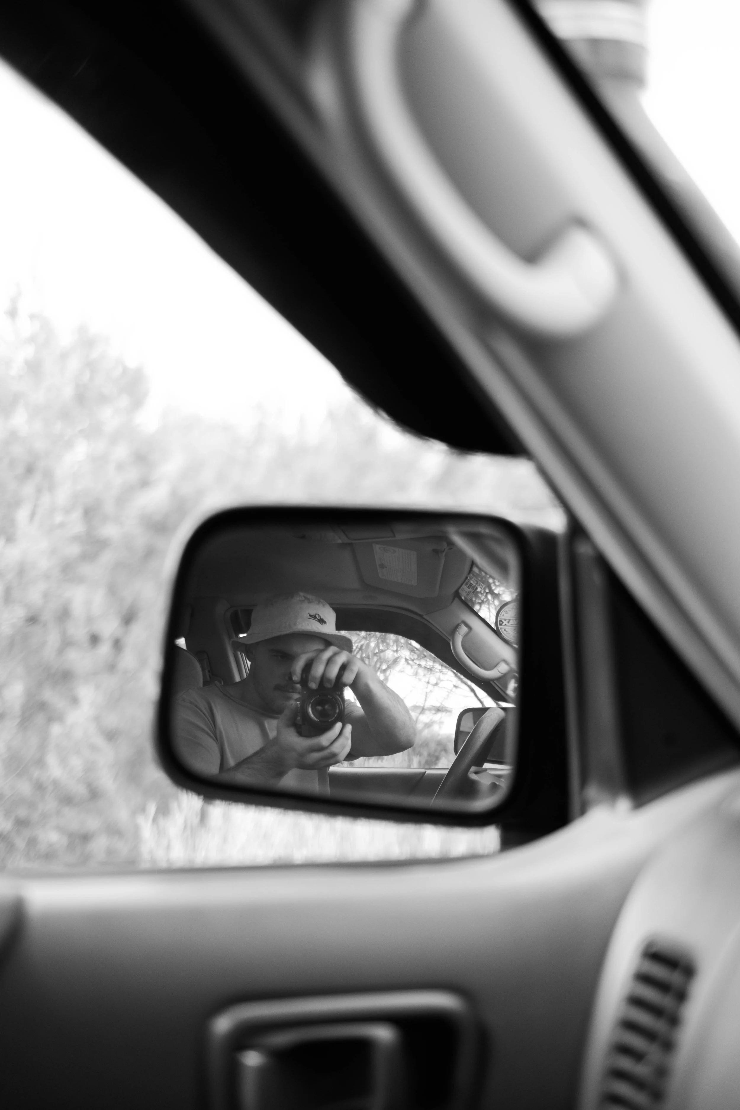 A person wearing a hat is taking a photo of their reflection in a car's side mirror. The interior of the car is visible, and the background shows trees and an overcast sky.