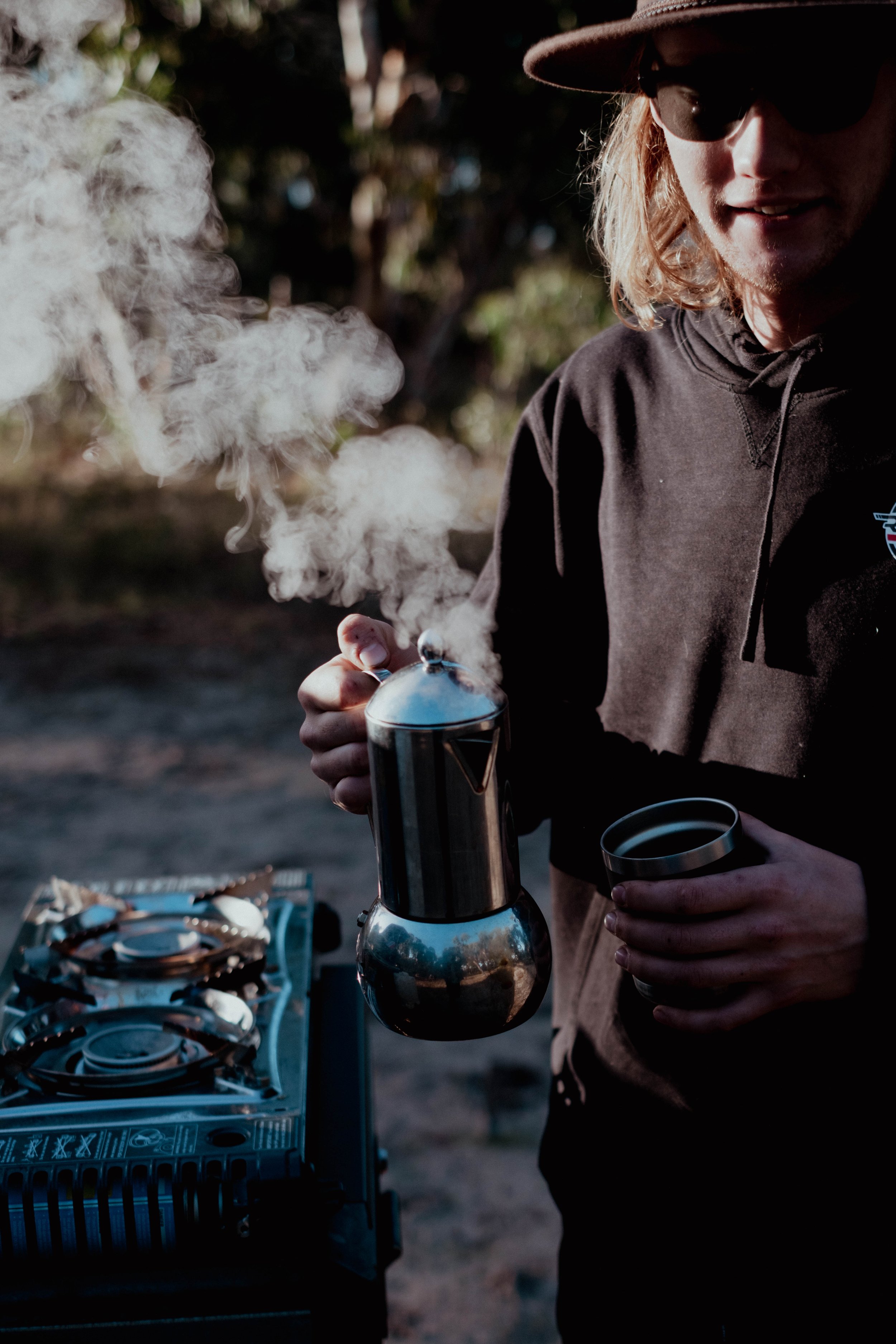 A person with long blond hair, wearing sunglasses, a hat, and a dark hoodie, is brewing coffee using a Chemex coffee maker outdoors during daytime. Steam rises from the brewing coffee, and a cup is held in the person's other hand. There is a tray of 