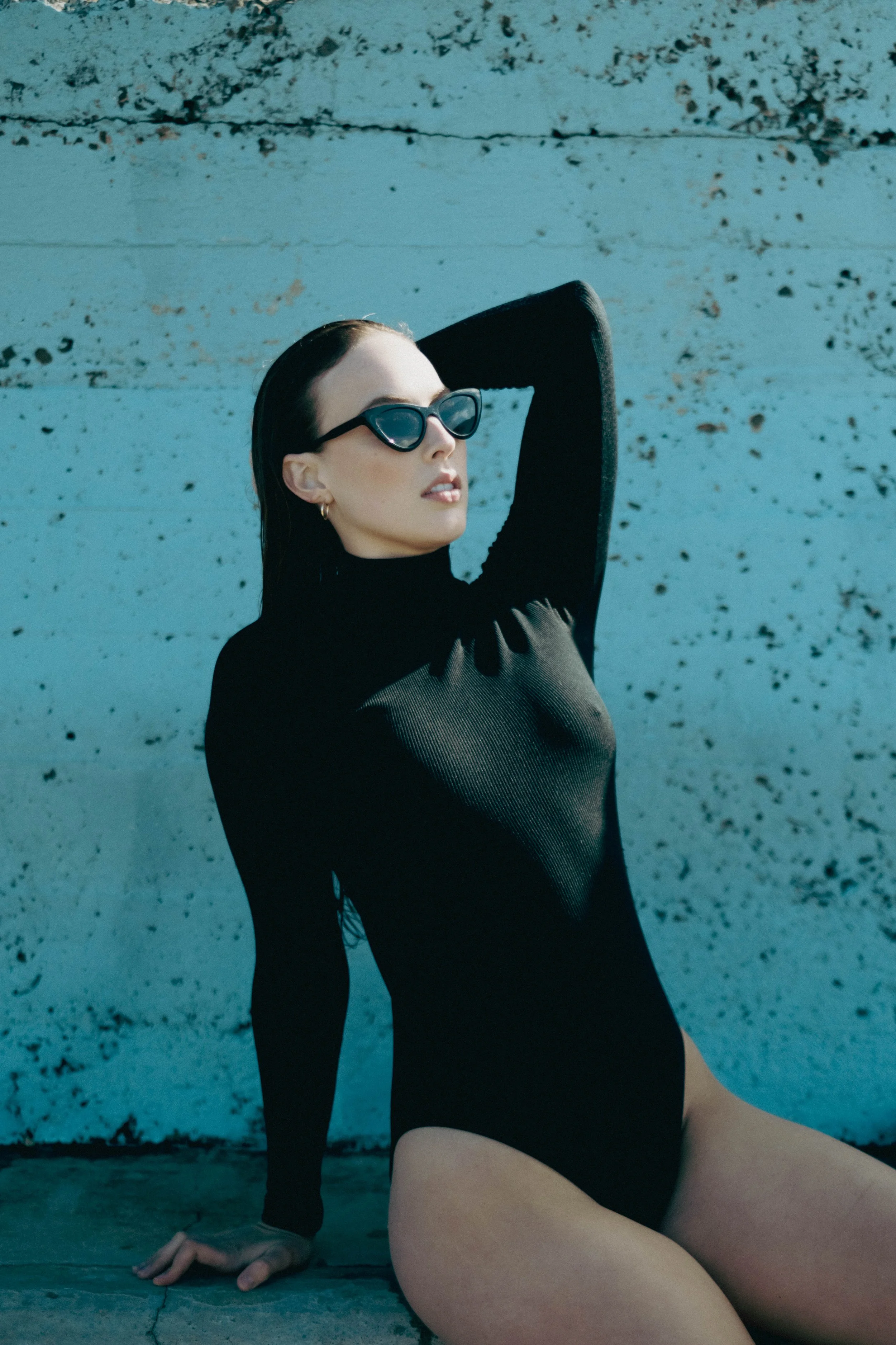 A woman with short dark hair wearing black sunglasses and a black long-sleeve top, posing with her arm behind her head, sitting on the ground against a textured wall with peeling paint.