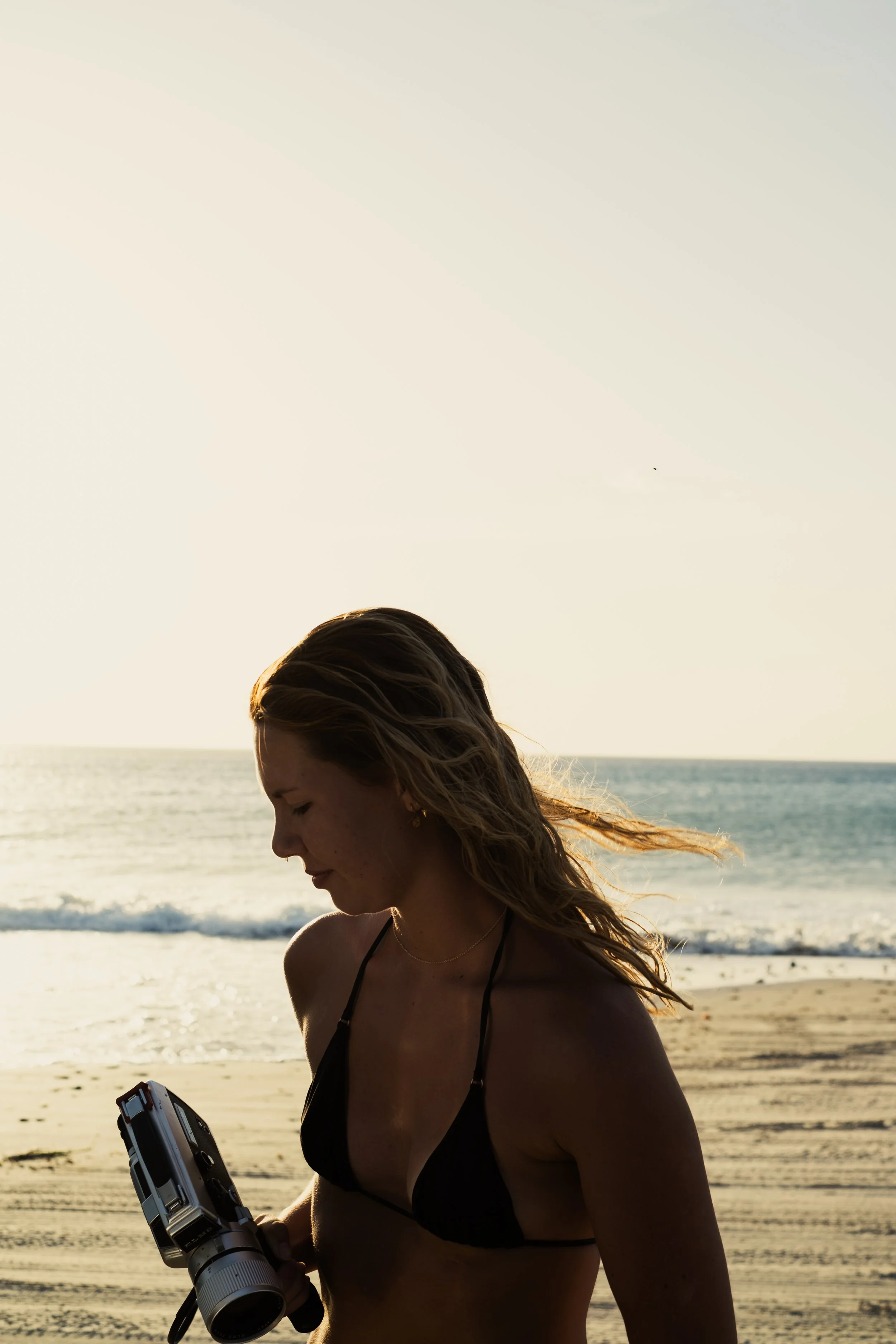 Young woman in a black bikini on a beach, holding a camera, with the ocean and sunset in the background.