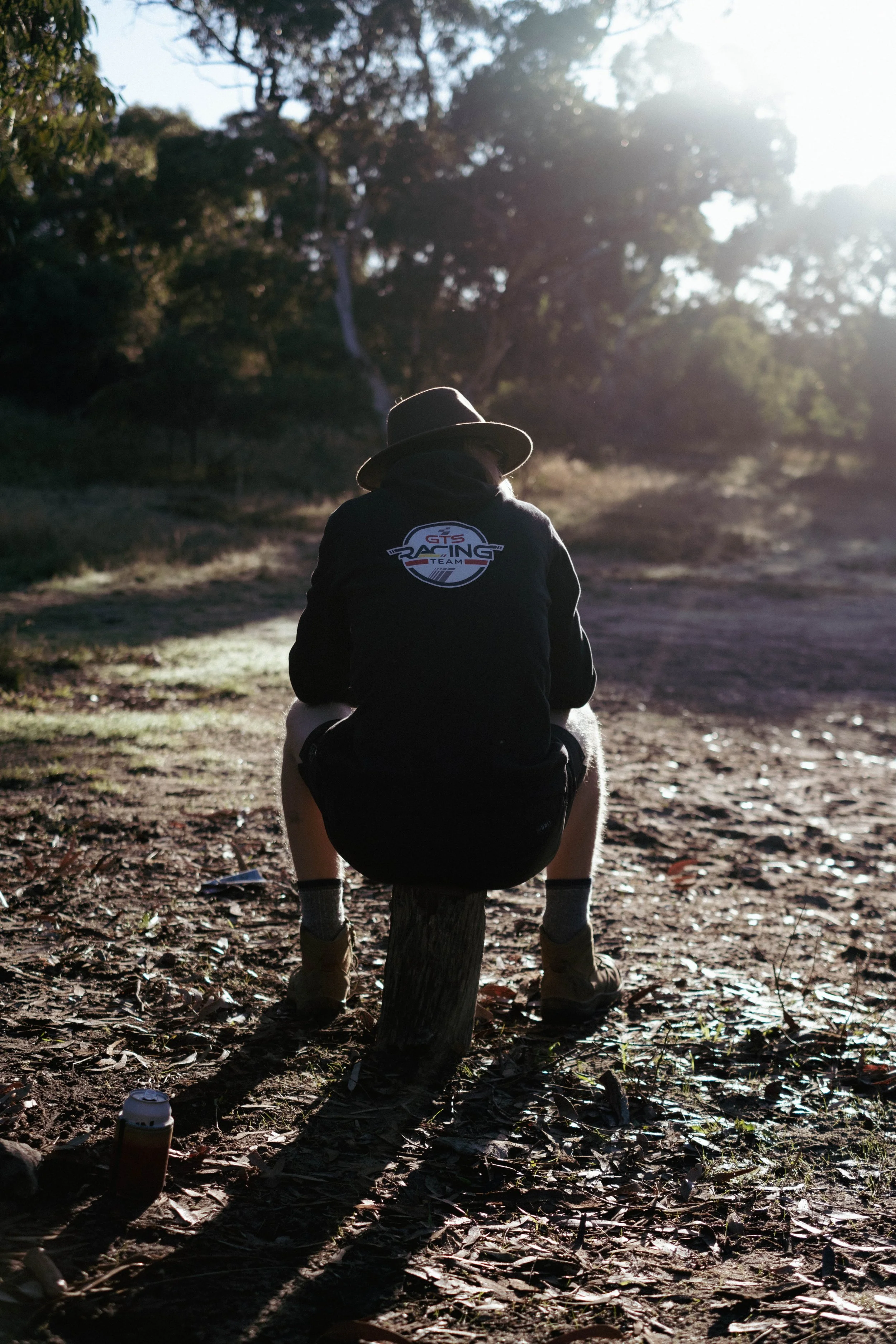 Person wearing a hat and black hoodie sitting on a small tree stump in a wooded area with sunlight filtering through trees.