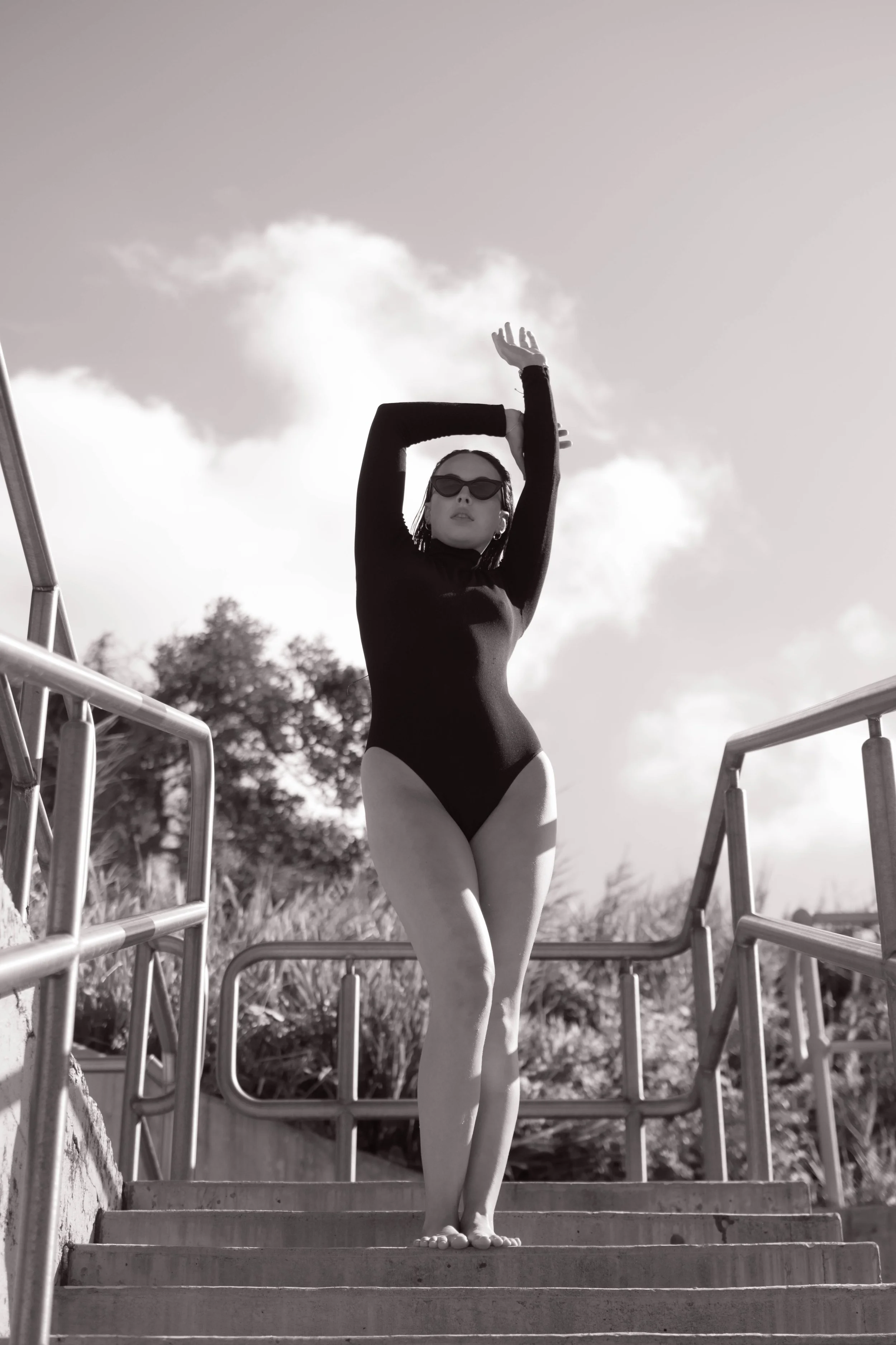 A woman with sunglasses, wearing a long-sleeve black swimsuit, stands outdoors on stairs with hand behind head and other arm raised, against a backdrop of trees and a cloudy sky.
