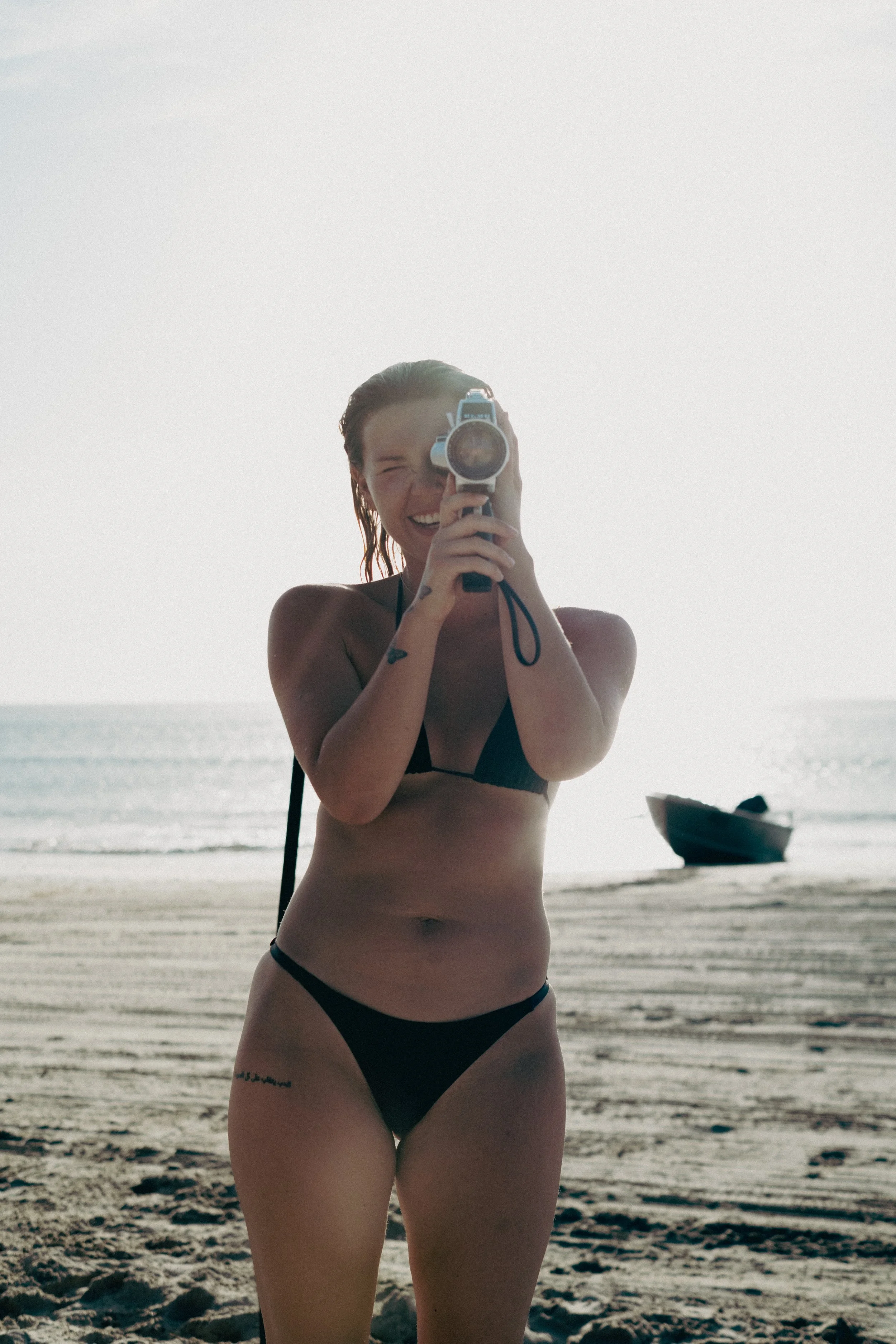 A woman in a black bikini taking a photo with a camera on a beach during sunset, with a boat in the background.