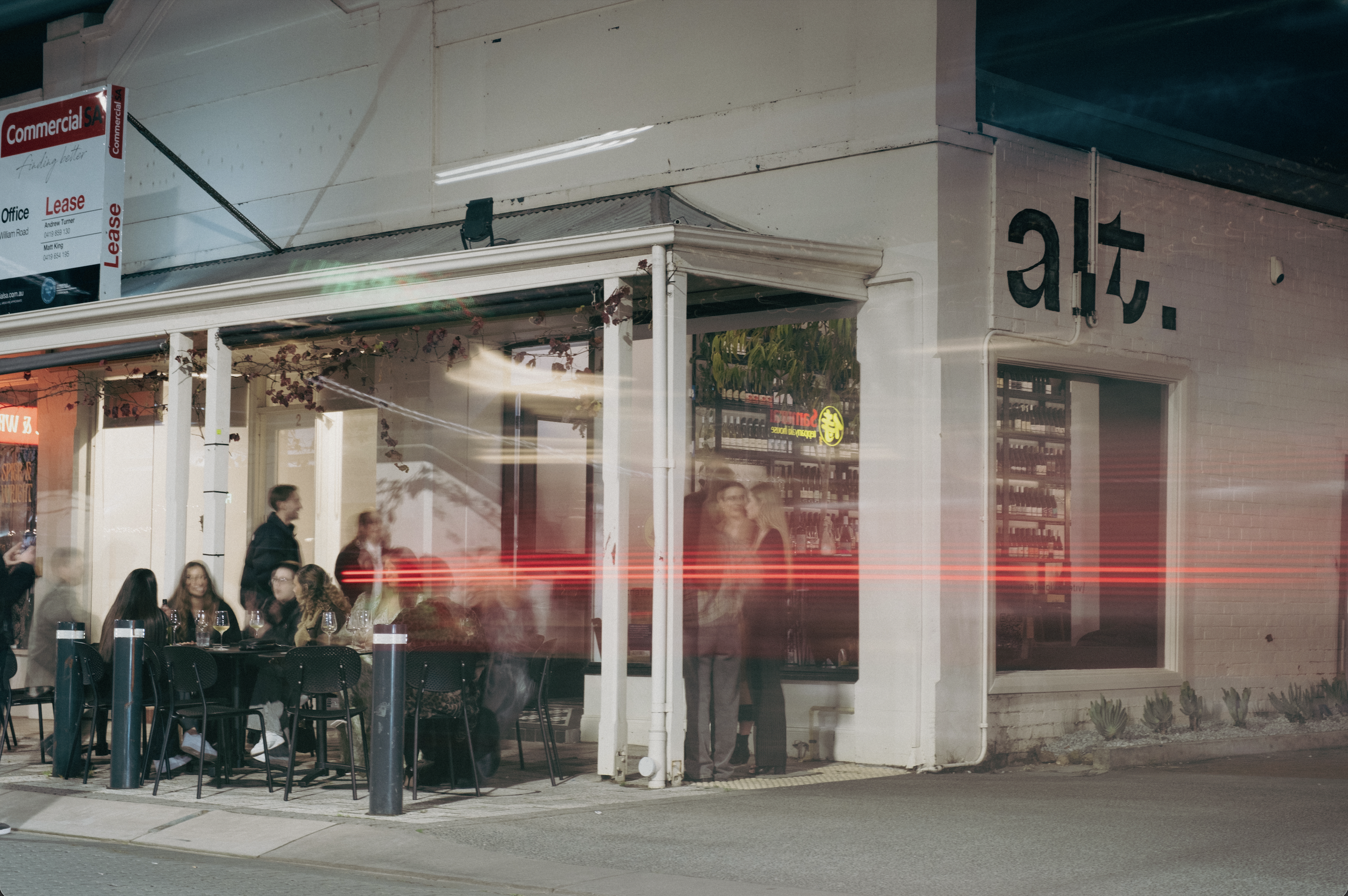 Nighttime street scene of a storefront with people sitting at tables outside. Light streaks from passing traffic are visible in front of the scene.