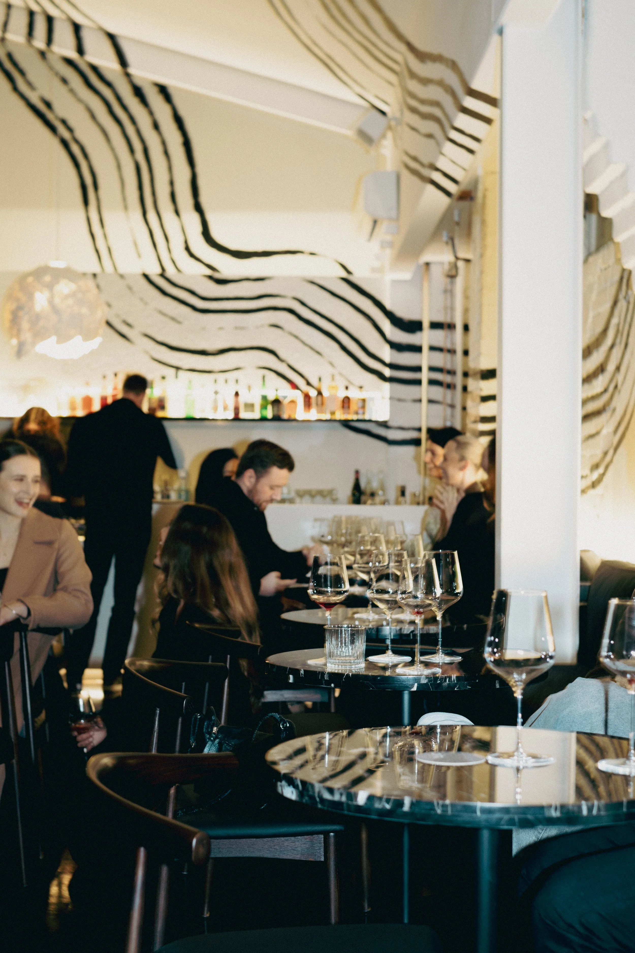 A restaurant scene with people dining, including a woman in a tan blazer and woman in black, under a zebra-striped decorative wall. There are wine glasses on the tables and a bar with bottles in the background.