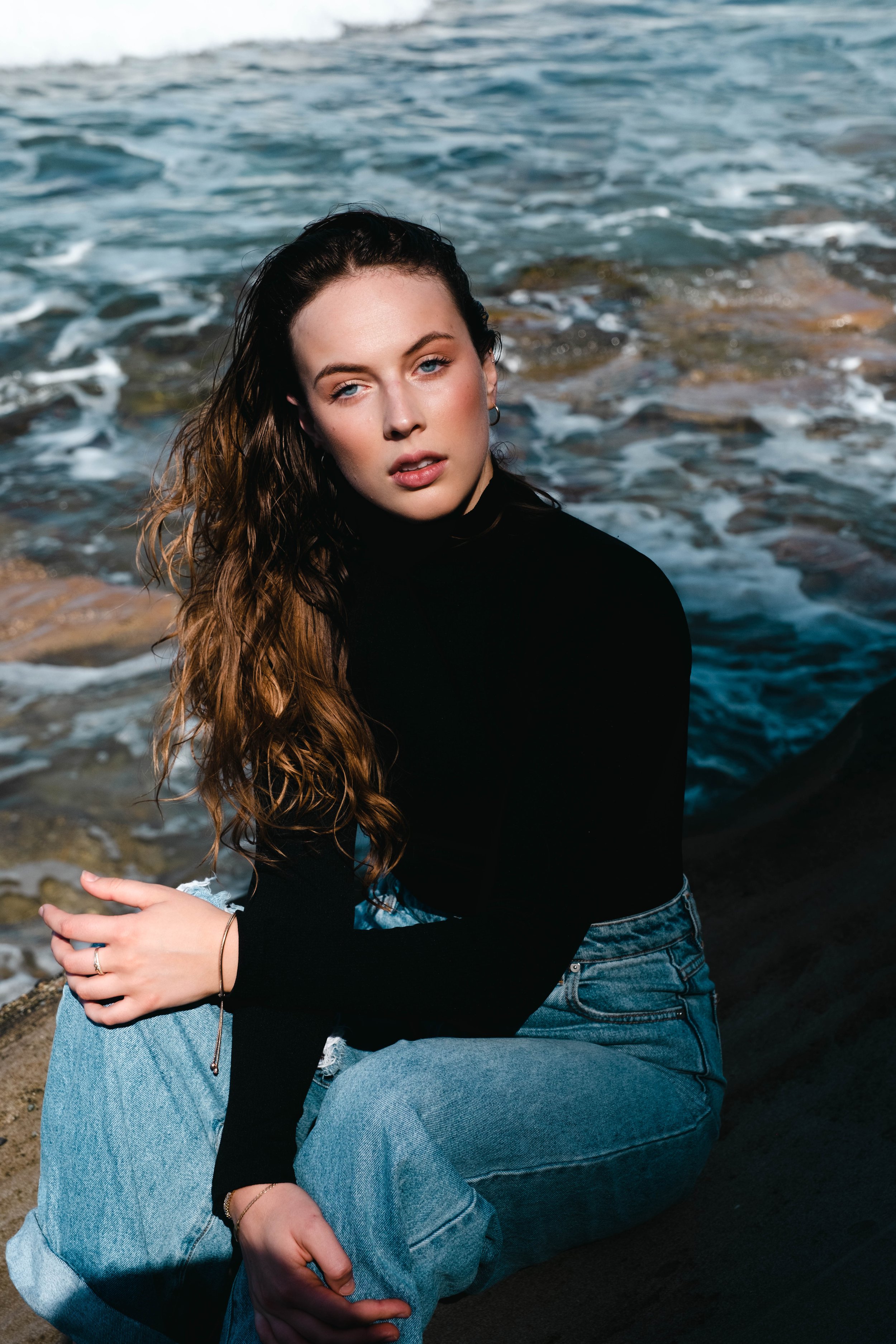 A woman sitting on the rocky shoreline with ocean waves in the background, wearing a black top, blue jeans, and jewelry.