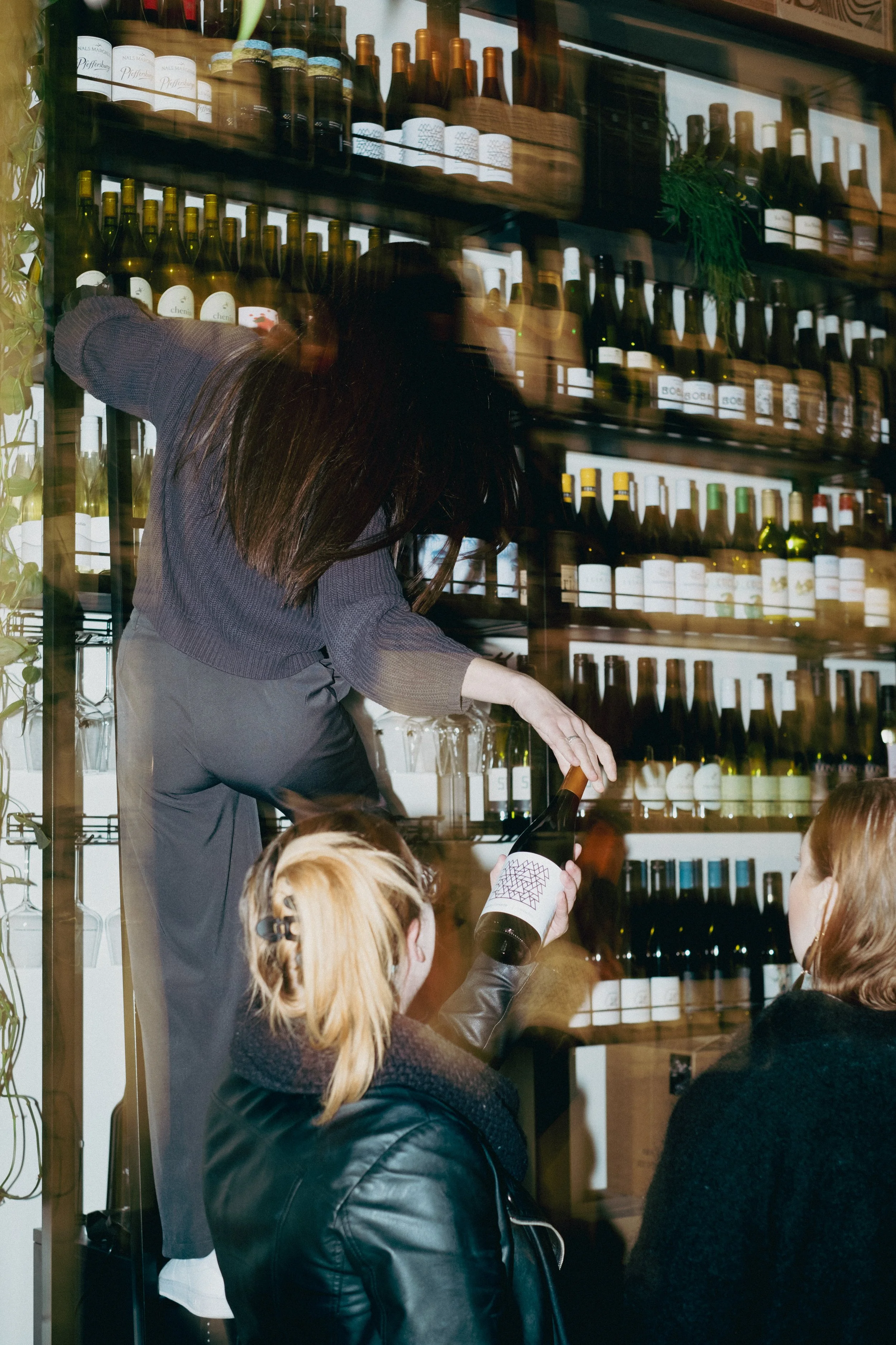 A woman with long dark hair reaching into a wine bottle on a shelf behind a glass window. Several children in the foreground, one girl with blonde hair in a ponytail with a bow. The scene appears to be a wine tasting event or a similar gathering.