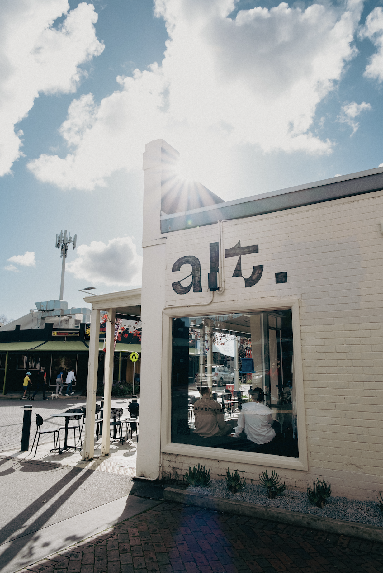 A cafe with large window seating, people inside and outside, and a sign with partially visible text on the building's exterior.
