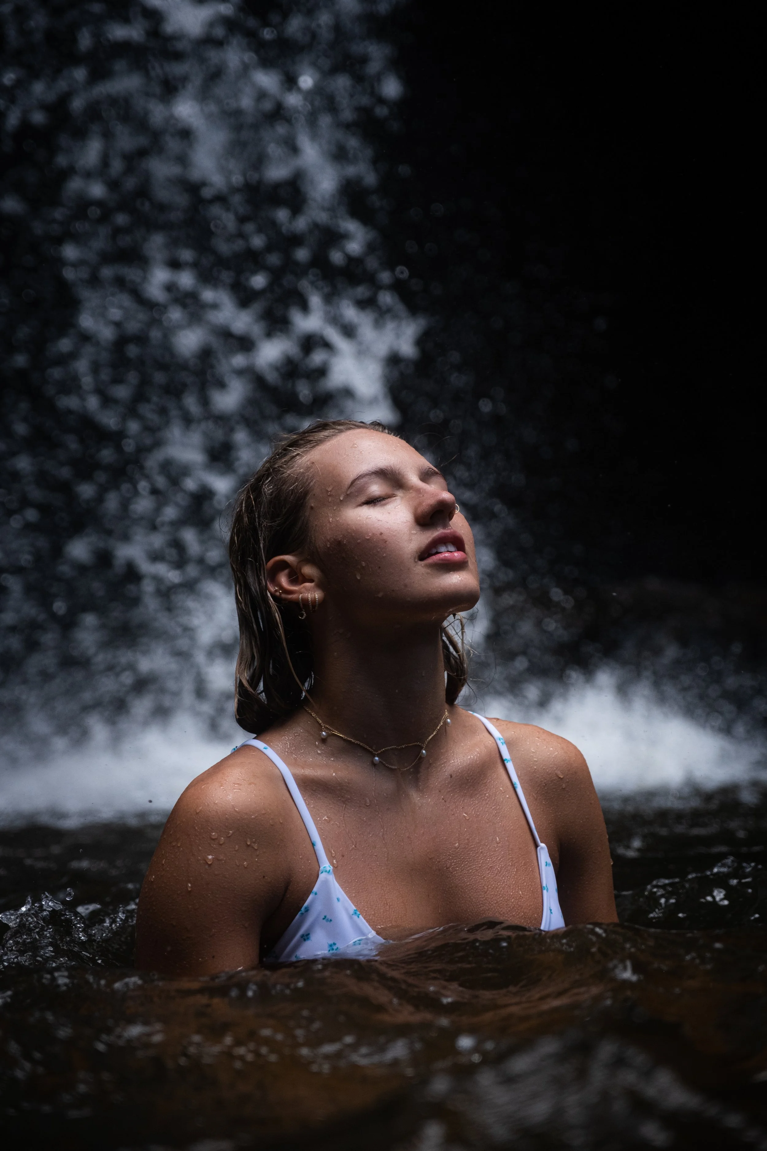 A woman with wet hair and closed eyes wears a white bikini with floral pattern, and layered necklaces, standing in water with a waterfall in the background.