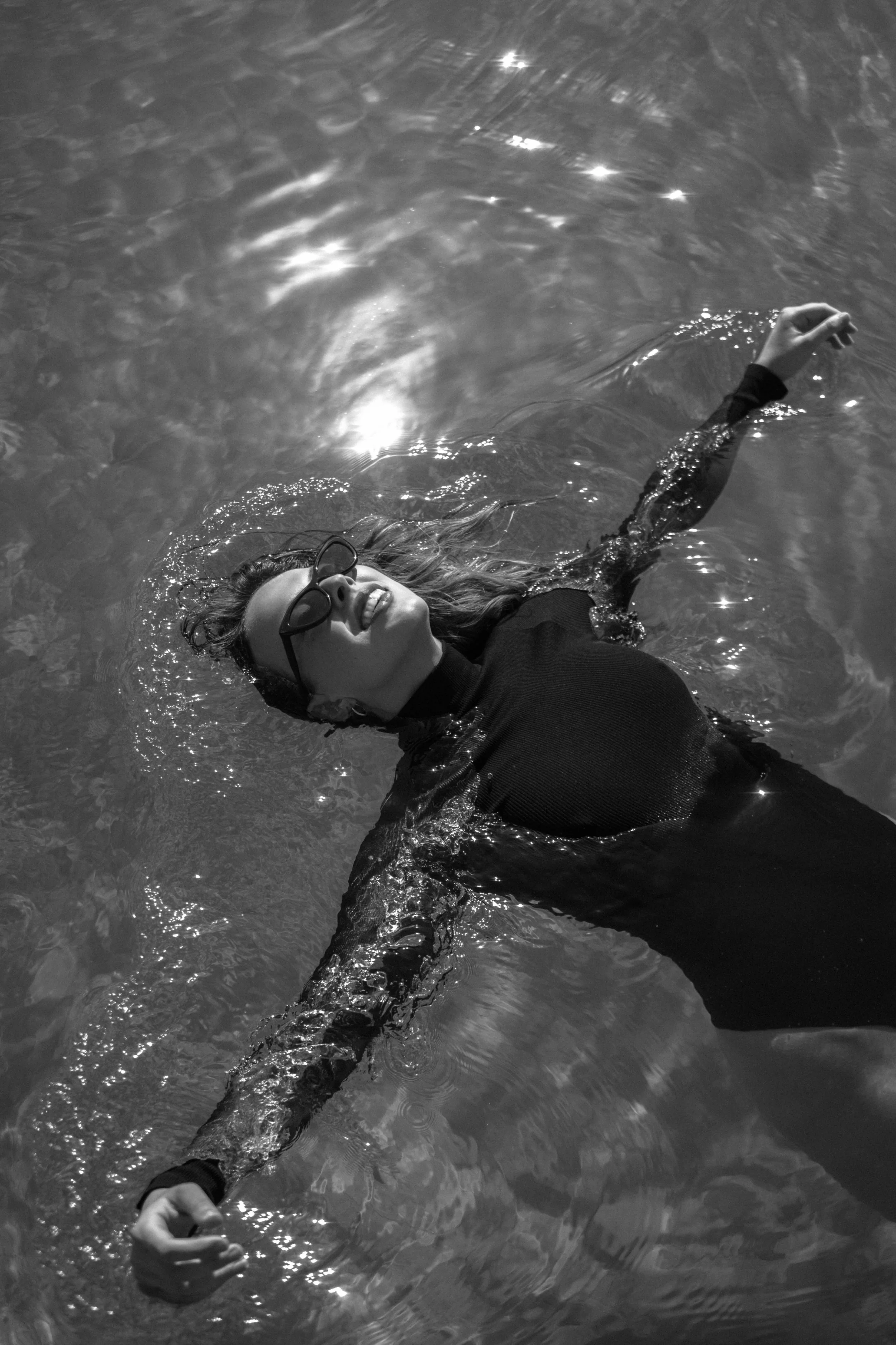 A woman wearing glasses and a black wetsuit is floating on her back in a body of water, smiling and looking up. Her hair is wet and spread out in the water, and her right arm is extended outward.