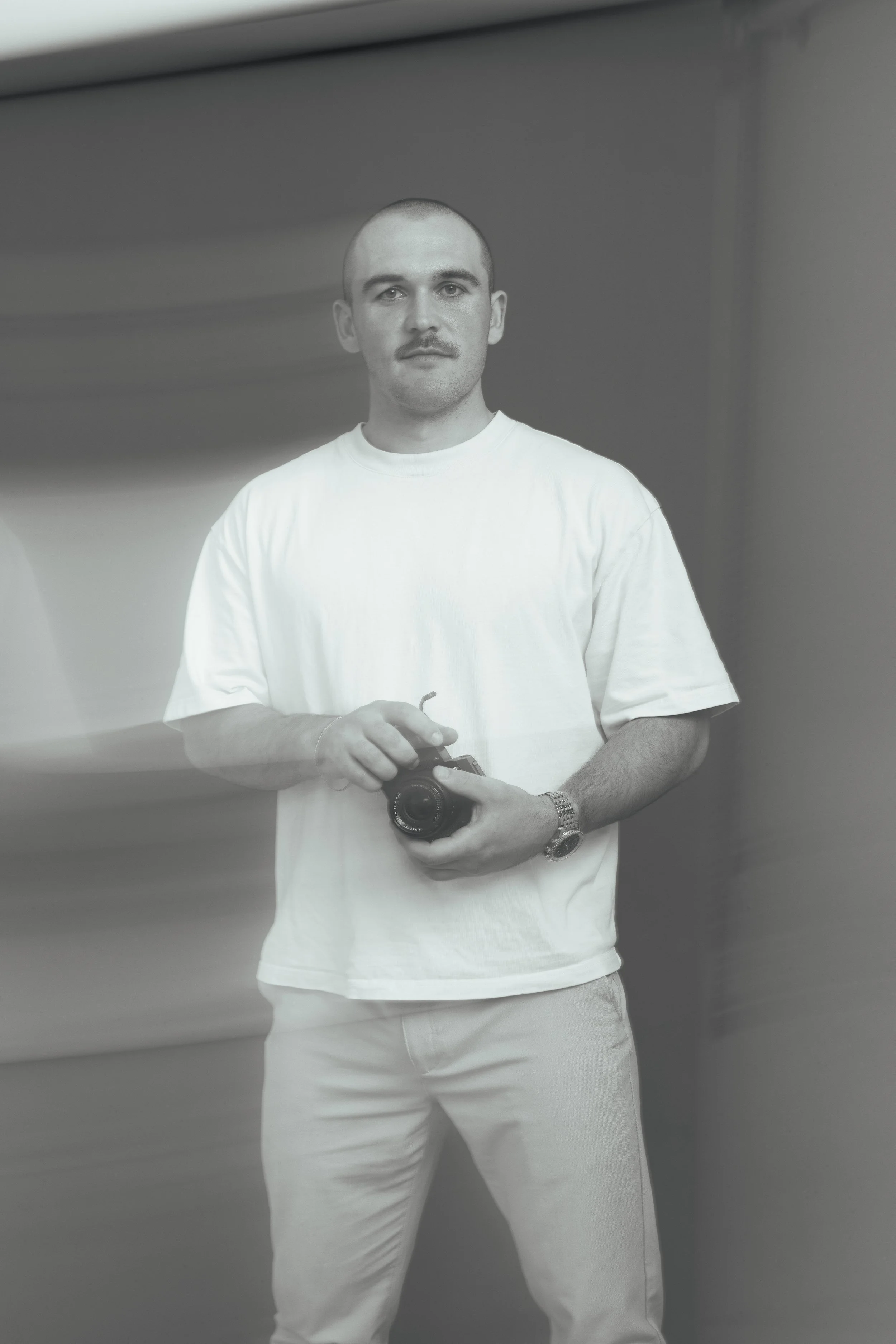 A man with a shaved head and a mustache wears a white t-shirt and holds a camera in front of a plain wall, looking directly at the camera.