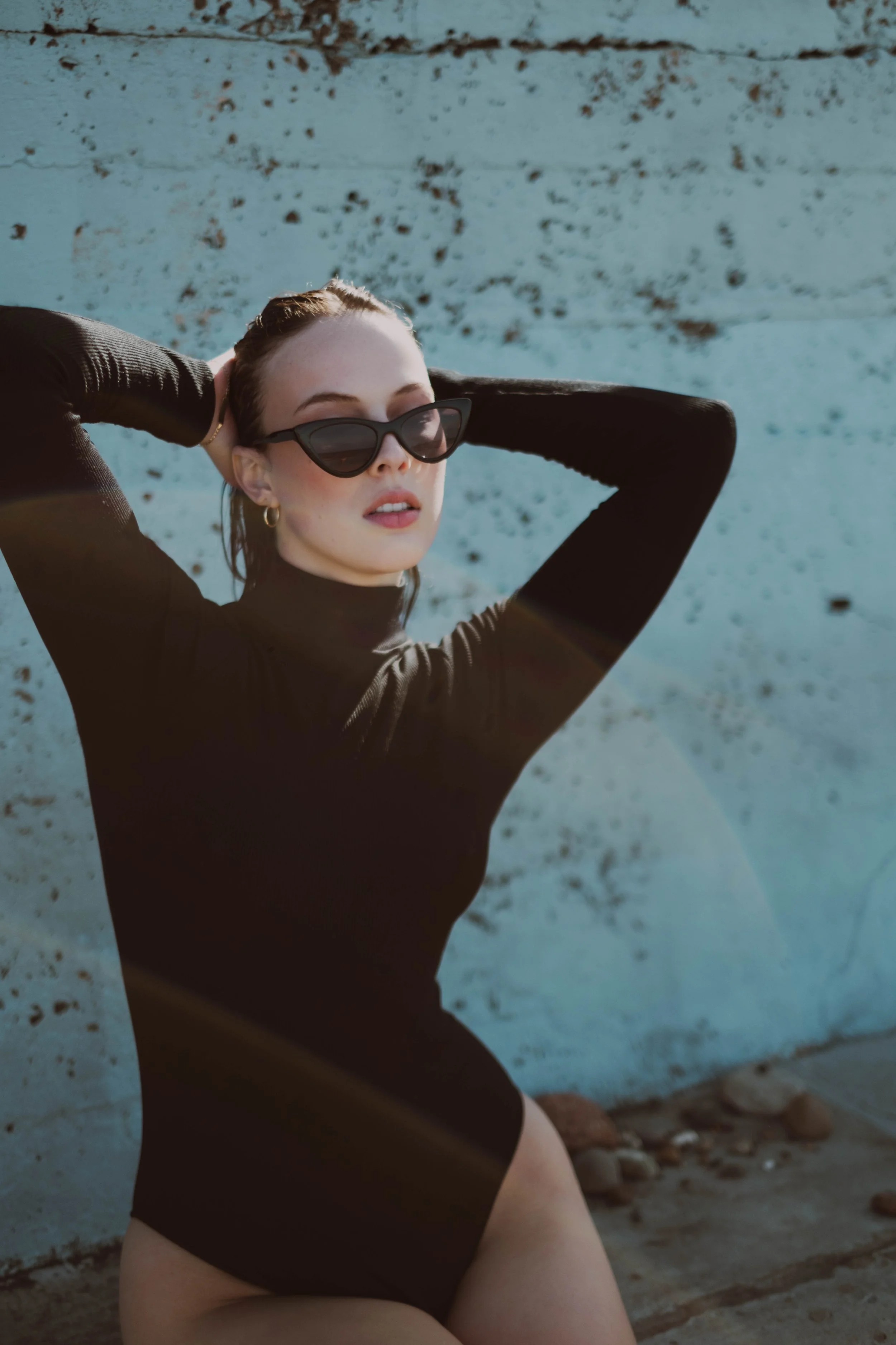 A young woman wearing black sunglasses and a black long-sleeve top, sitting with her hands behind her head against a weathered blue wall, with rocks on the ground.