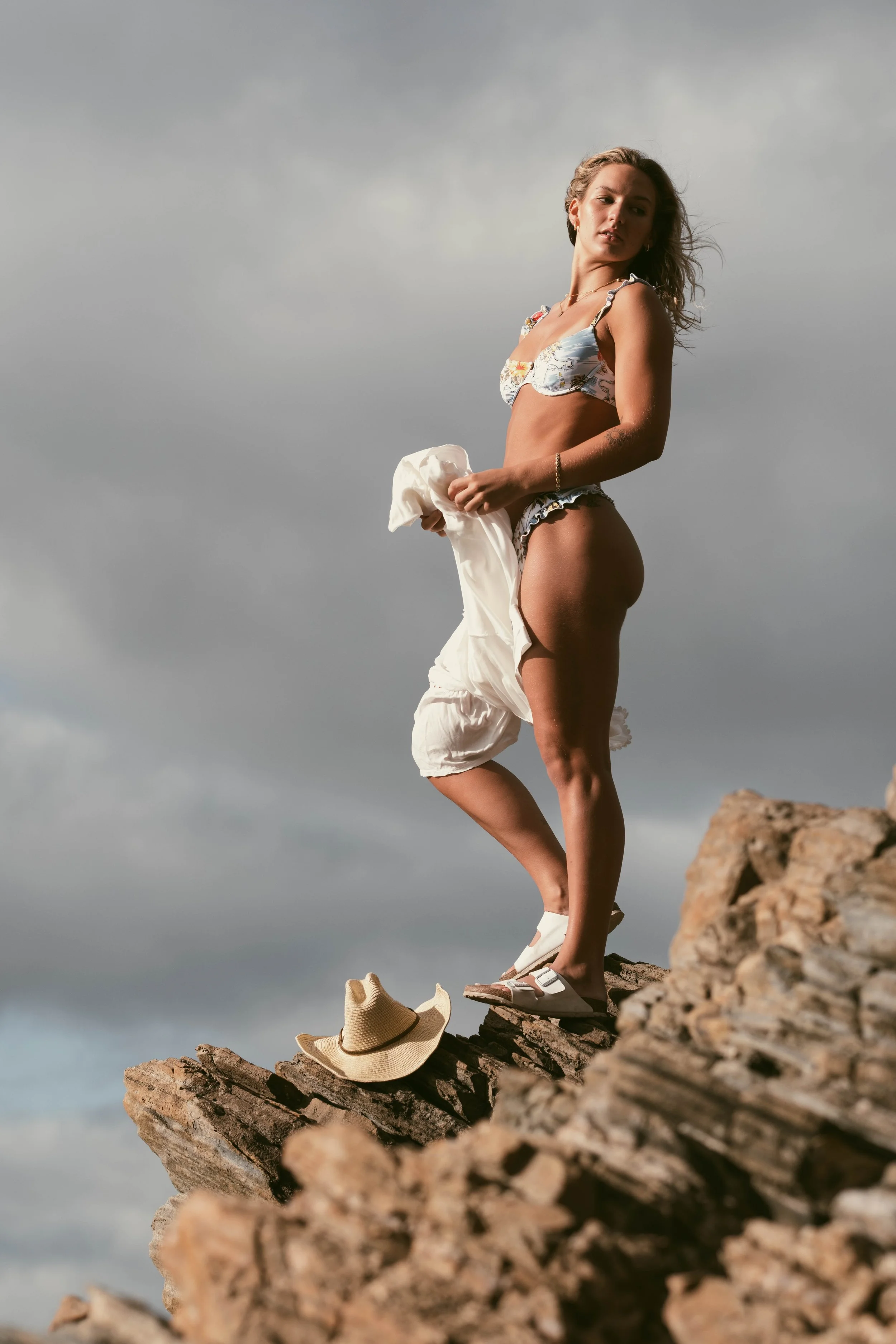A woman in a swimsuit stands on rocks at the beach, holding a white towel, with a sun hat and sandals nearby, under a cloudy sky.