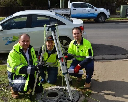 Three men wearing high vis jackets crouched down beside a tripod and some pipeline technology.