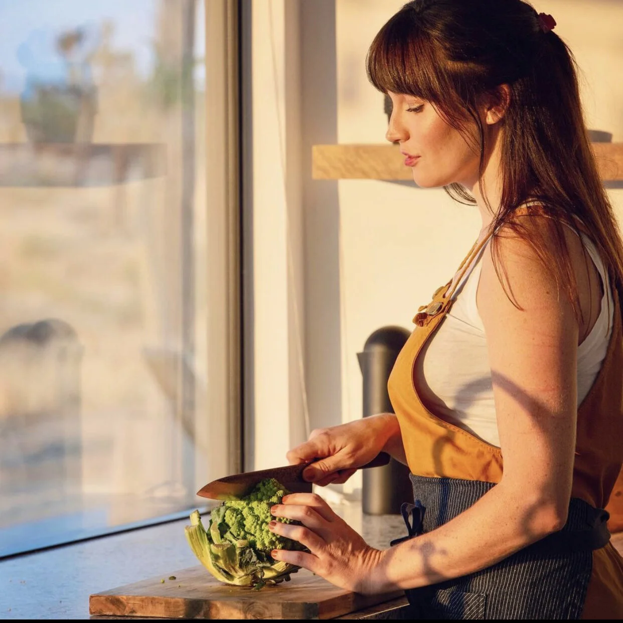 A woman with long brown hair in a ponytail, wearing a white tank top and a light brown apron, is cutting green broccoli with a black-handled knife at a kitchen counter near a window.