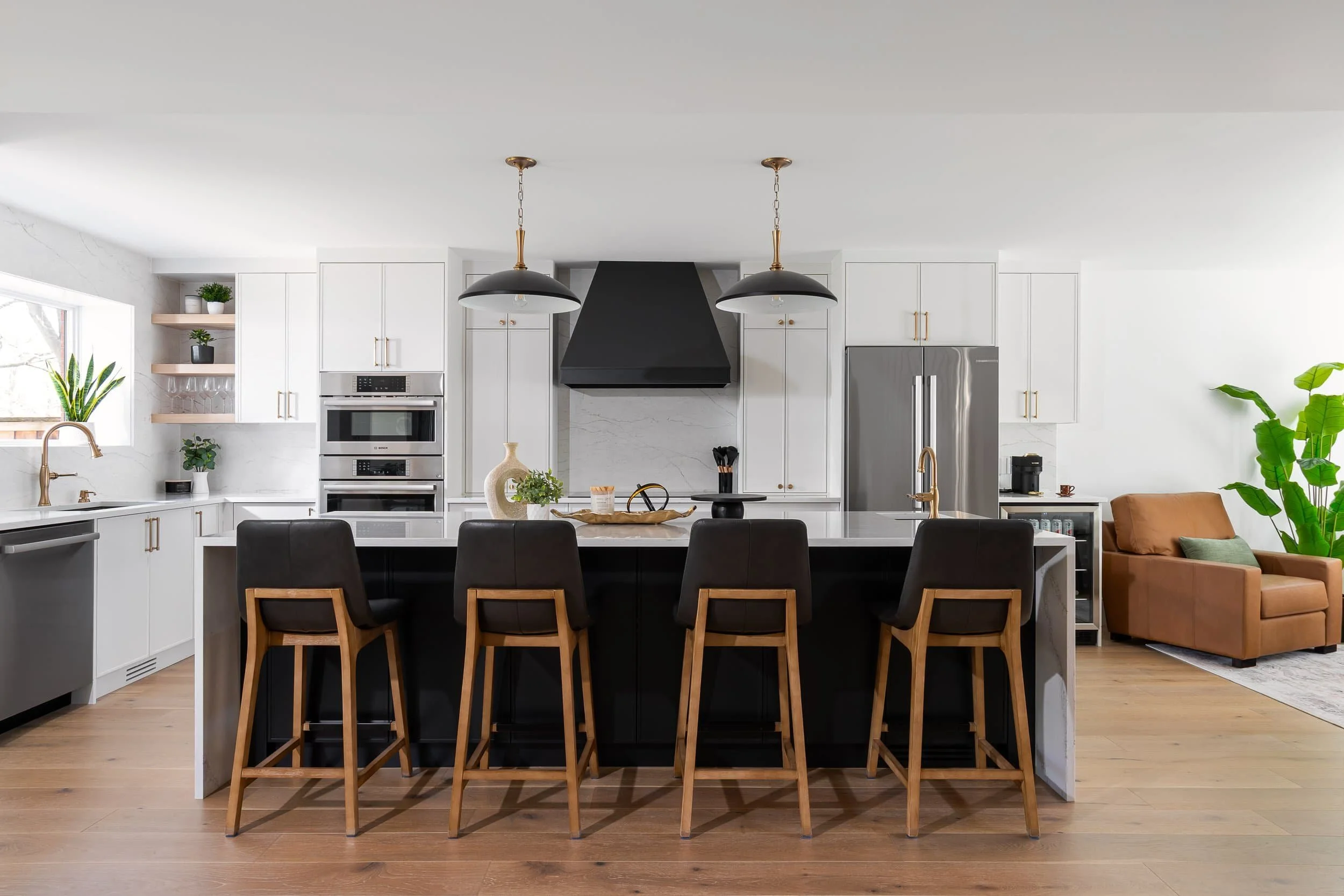 Modern kitchen with white cabinetry, a black island with four stools, stainless steel appliances, and a brown armchair with plants.