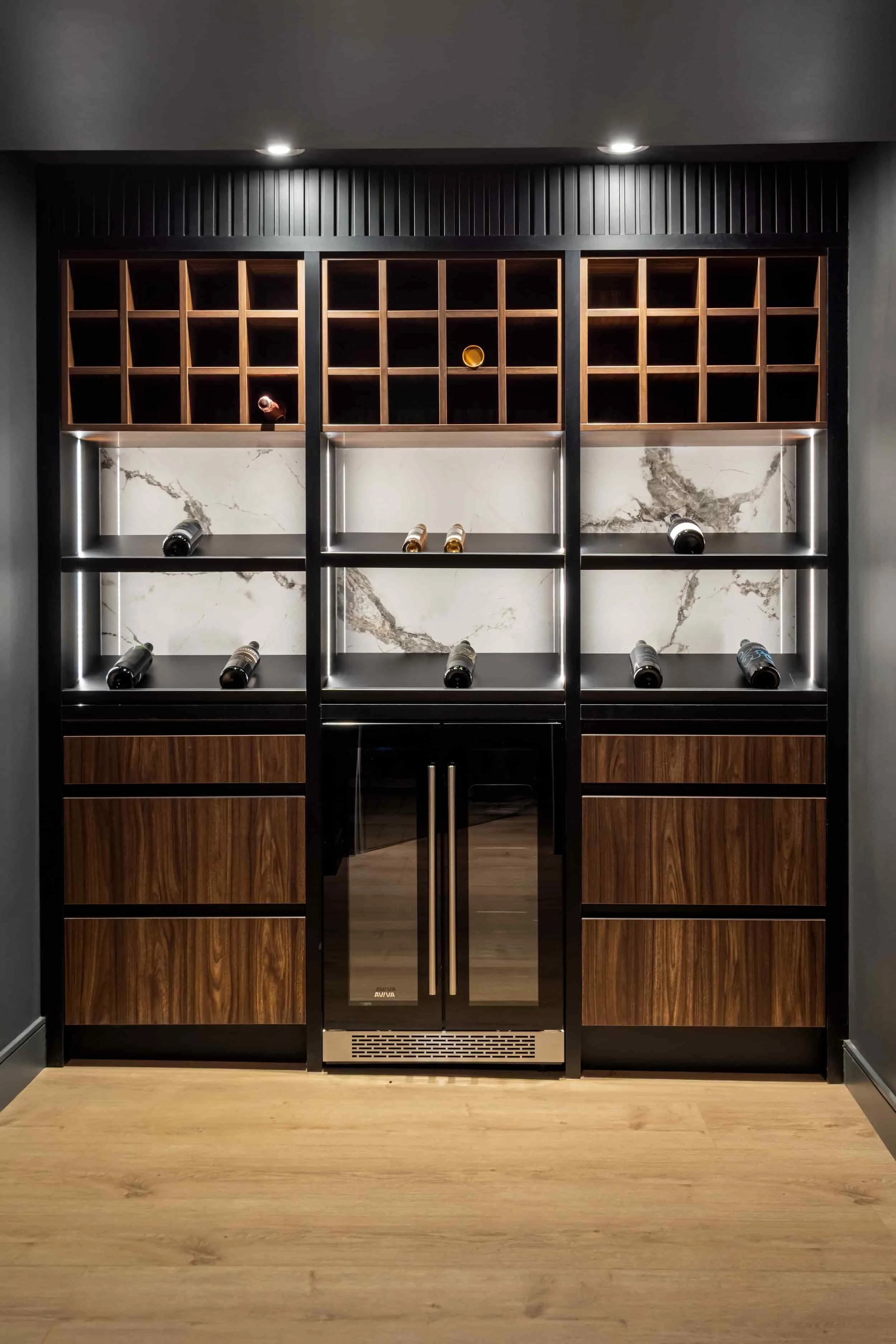 A wine storage unit with bottles and glasses, featuring wooden drawers, marble backing, and a small wine cellar in the centre.
