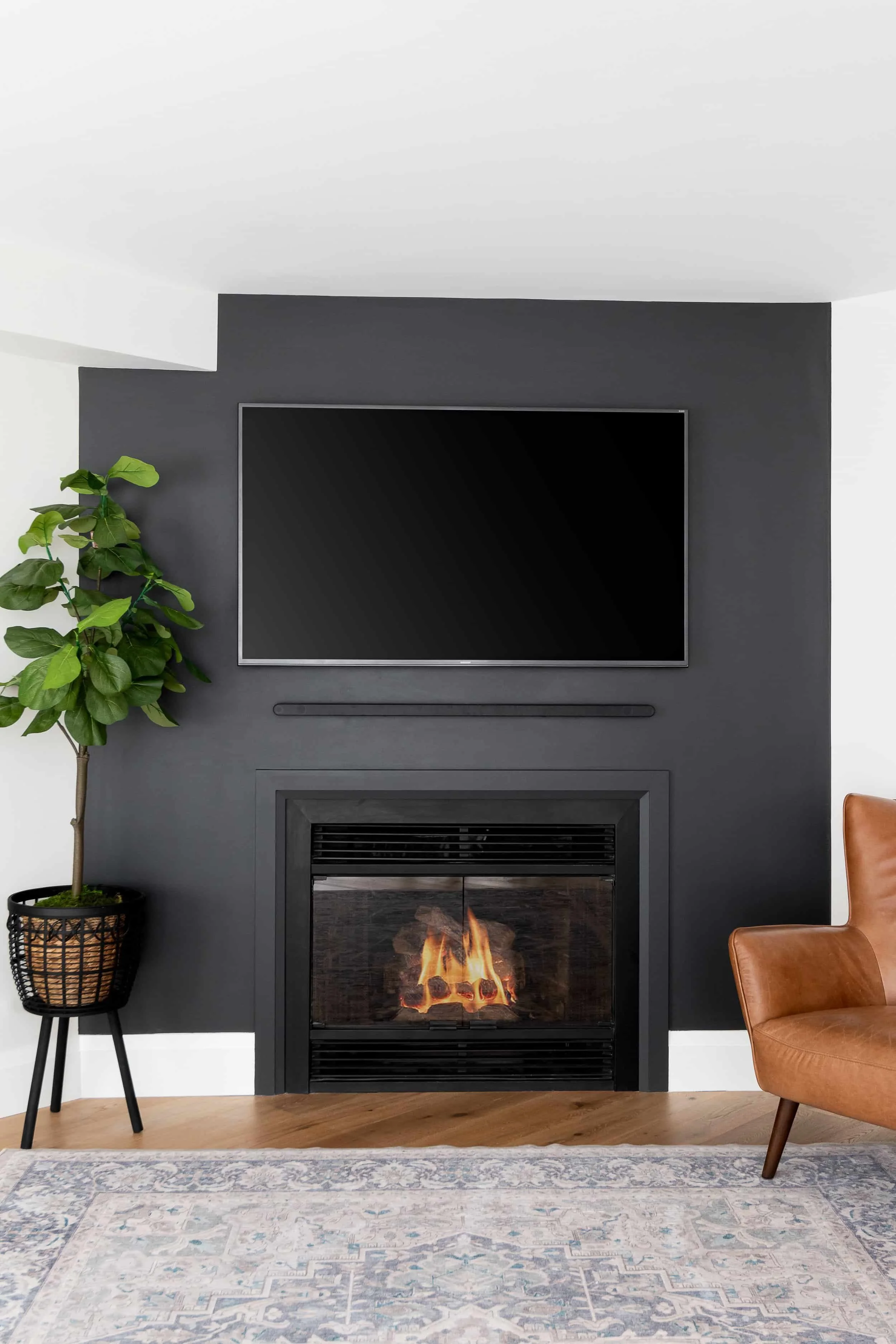 Living room with a black accent wall, mounted flat-screen TV, fireplace with flames, potted plant on the left, a tan leather chair on the right, and a patterned rug on wooden floor.
