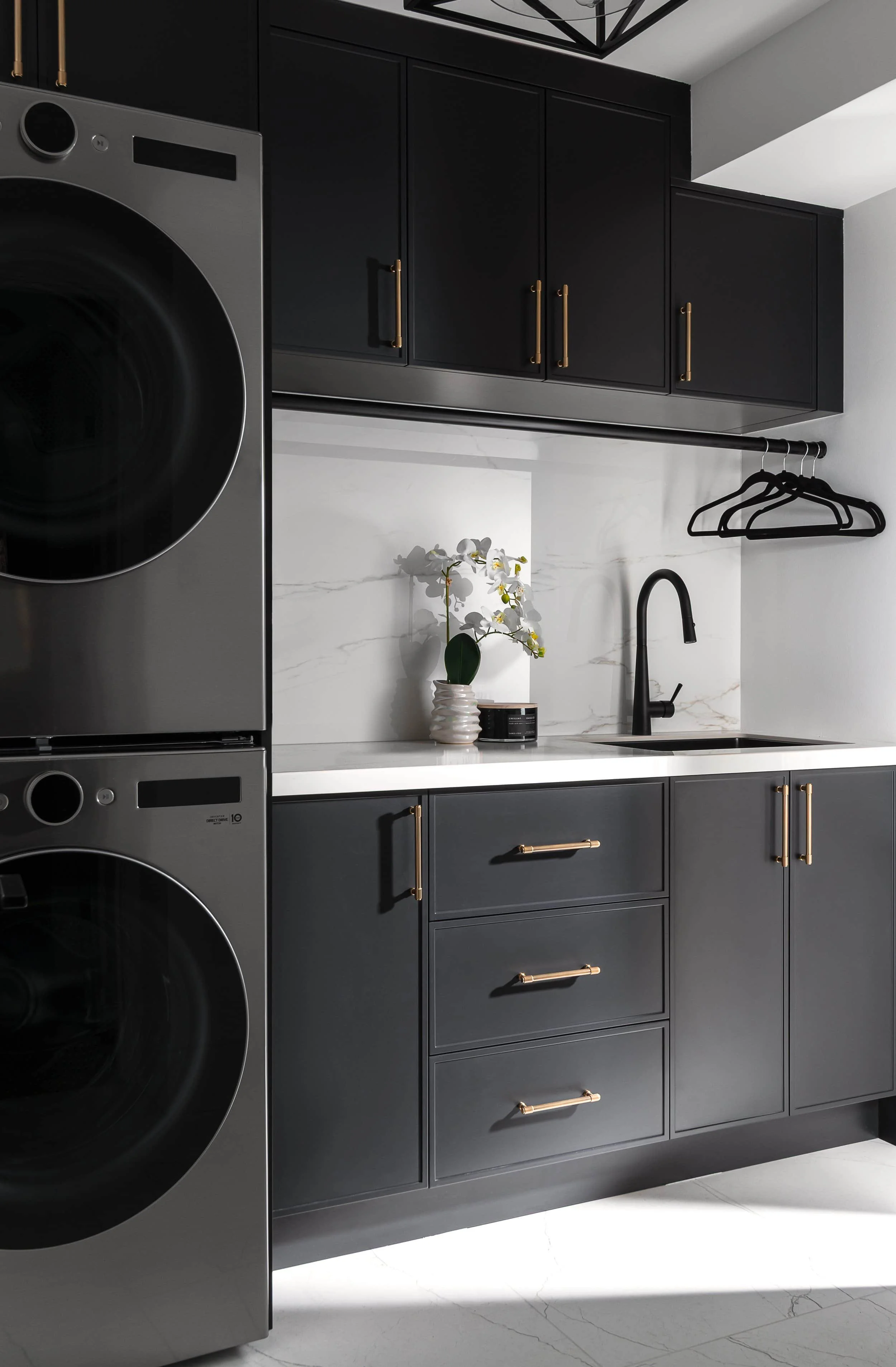 Modern laundry room with black cabinetry, white marble backsplash, stacked washing machine and dryer, black faucet, black hangers, and a small potted plant on the white countertop.