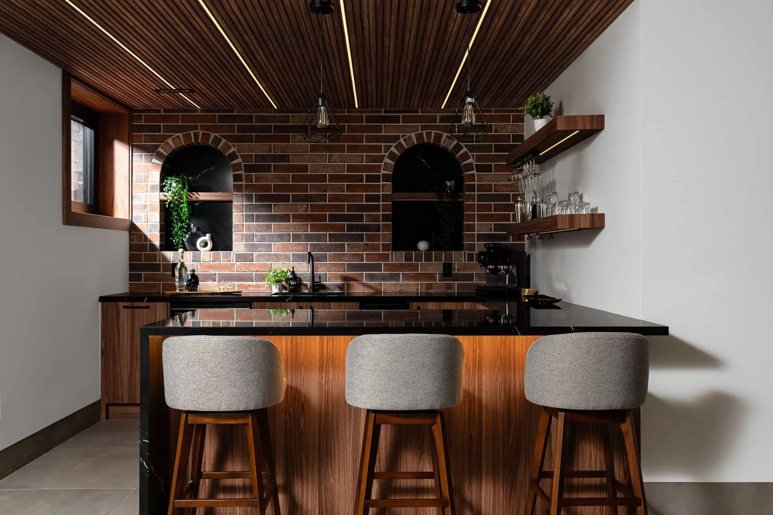 Modern kitchen with brick wall, black countertop, wooden accents, bar stools, and open shelving with glassware.