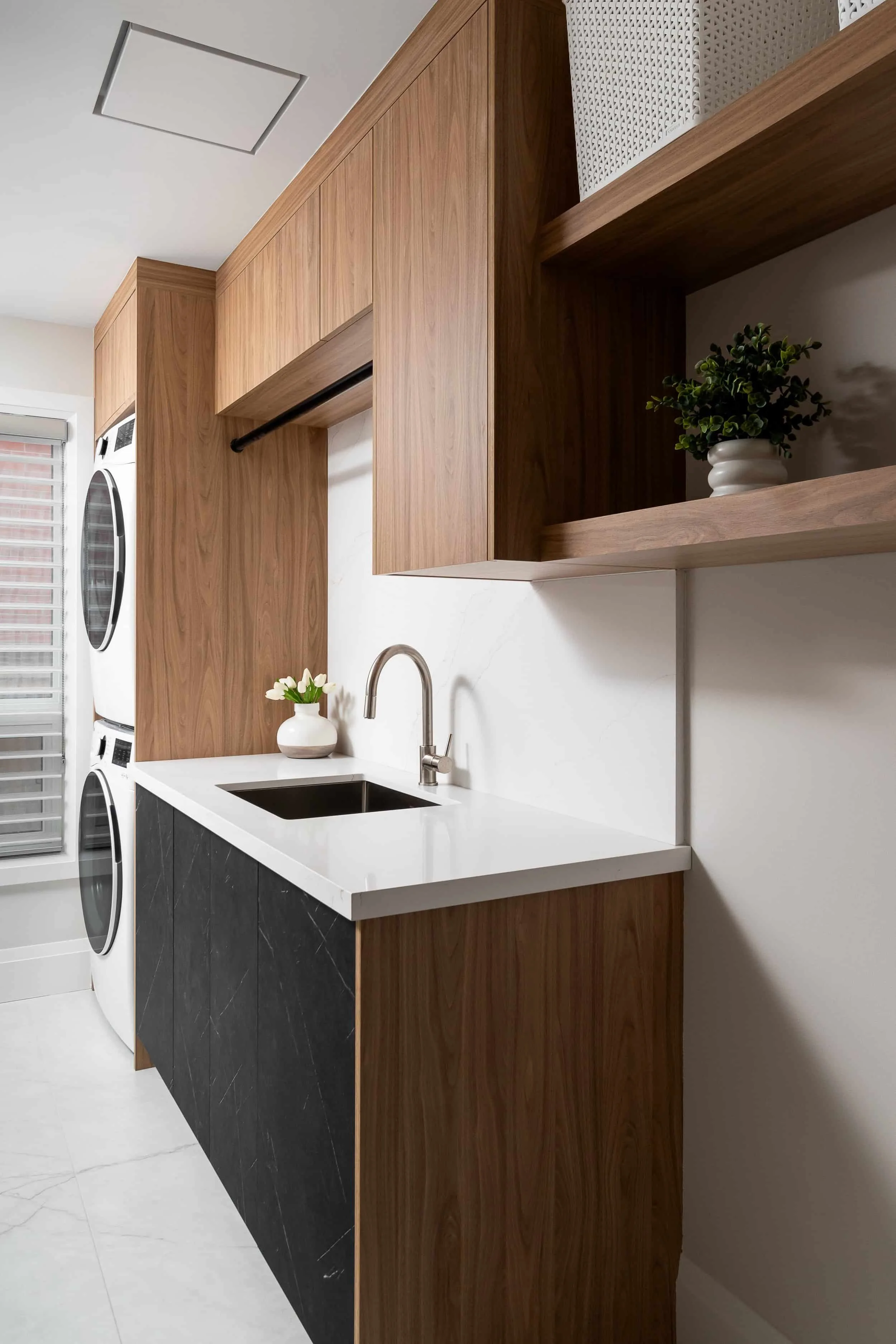 Modern laundry room with wooden cabinets, white countertop, small sink, and stacked washer and dryer, decorated with a small potted plant and a vase of white flowers.