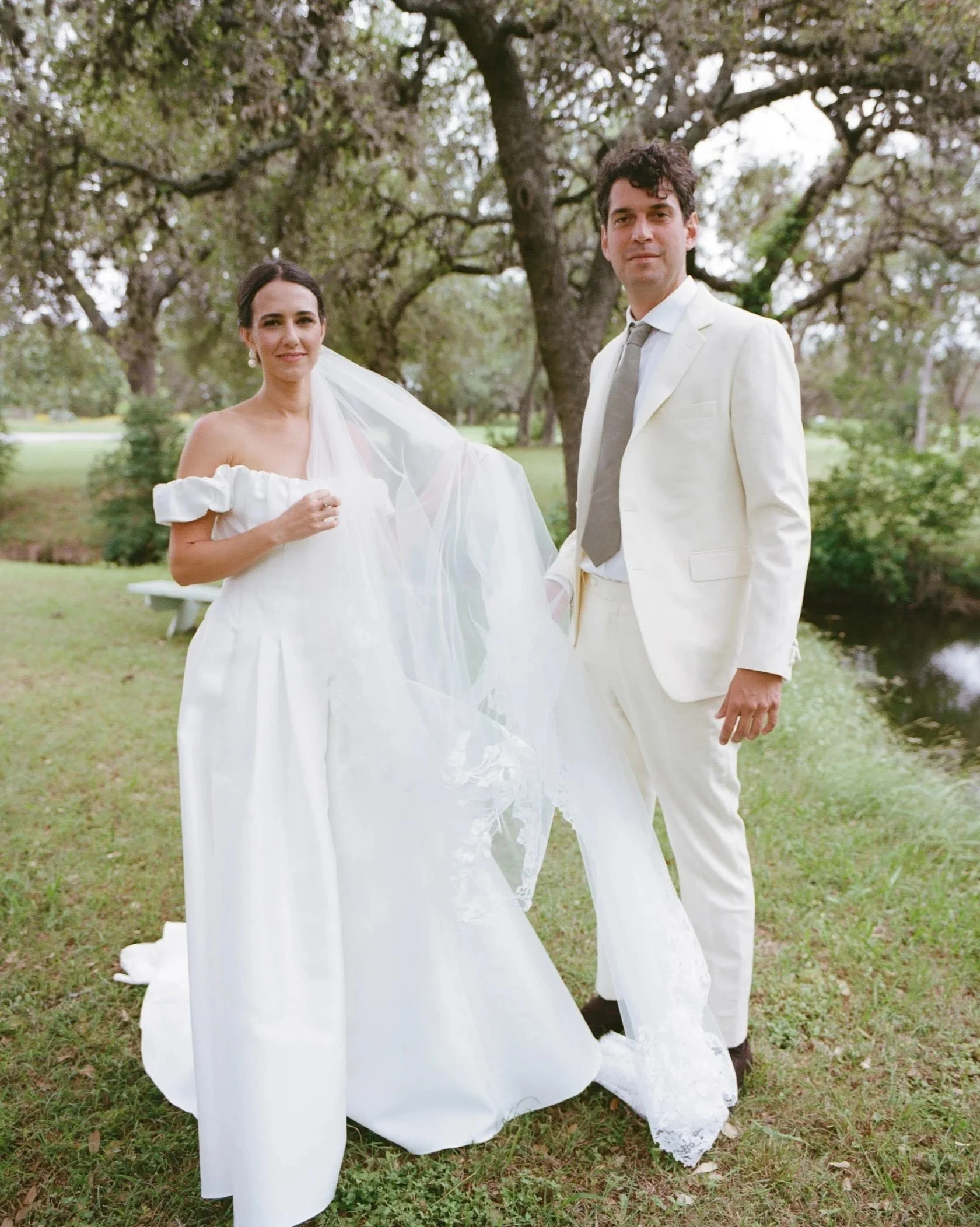 A bride and groom standing outdoors on grass with trees in the background. The bride wears a white off-shoulder wedding dress and veil, and the groom is in a white suit with a light gray tie.