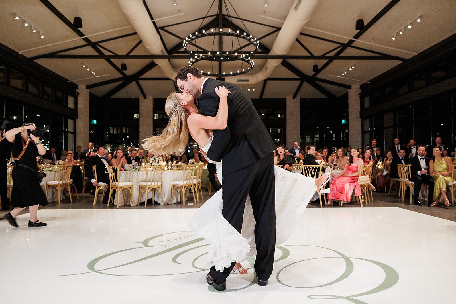 A newlywed couple dancing at their wedding reception in a ballroom, with guests seated at decorated tables watching and cheering.