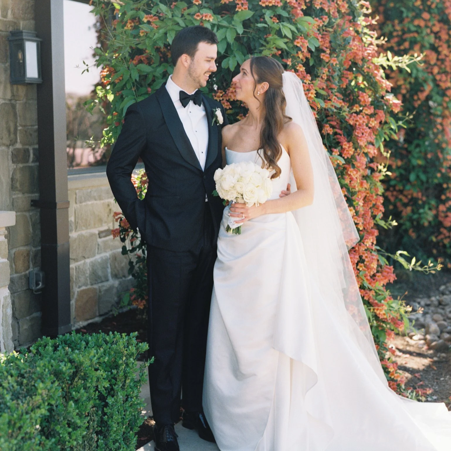 A bride and groom stand close together outdoors, smiling at each other. The groom is dressed in a black tuxedo with a bow tie, and the bride wears a white wedding gown with a veil and holds a bouquet of white roses. There are blooming orange and green plants behind them, and a stone wall to the left.