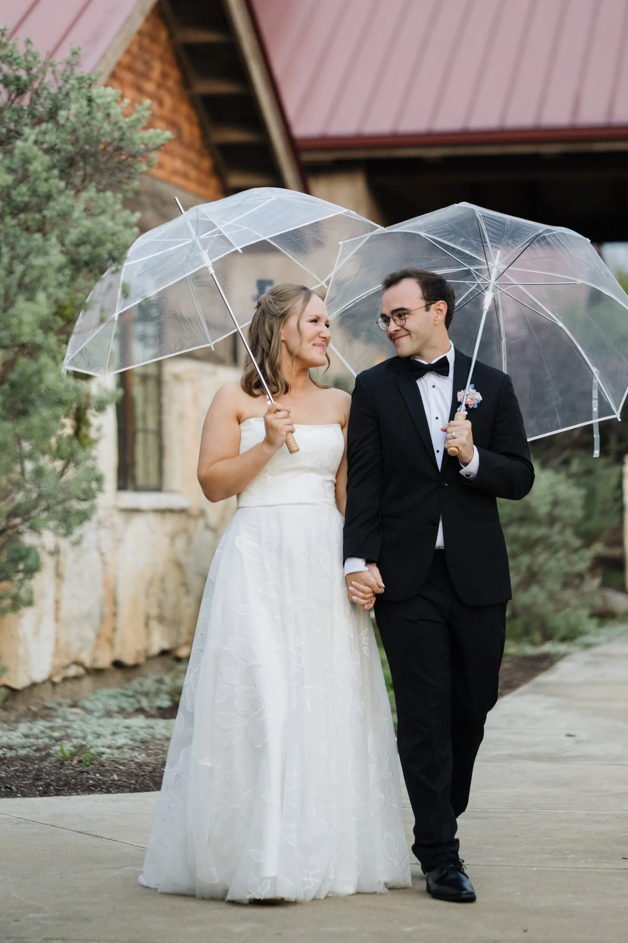 A bride and groom holding hands and sharing a moment under clear umbrellas outdoors, with a building and greenery in the background.