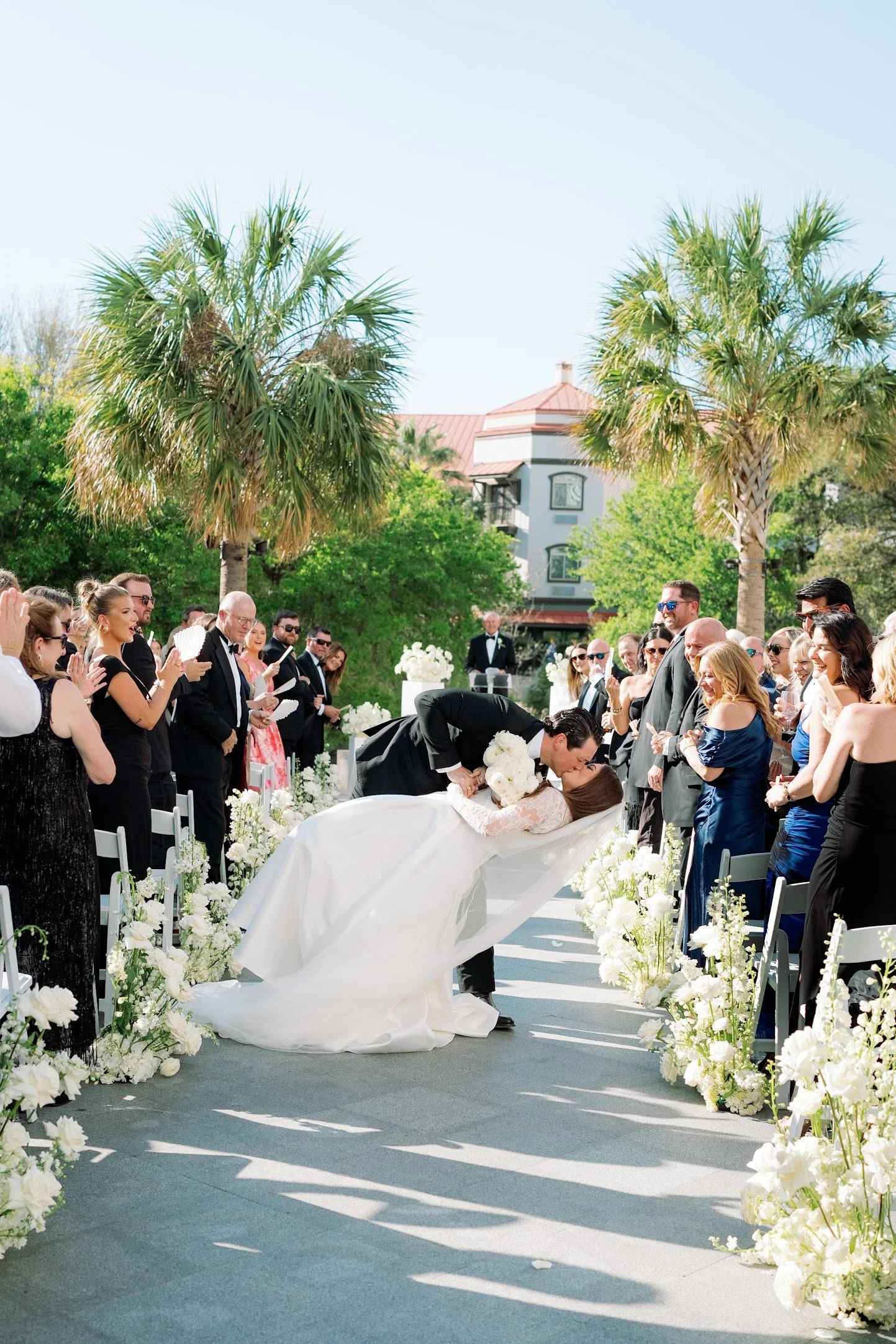 A bride and groom kiss during their outdoor wedding ceremony, surrounded by friends and family, with white floral decorations and two palm trees in the background.
