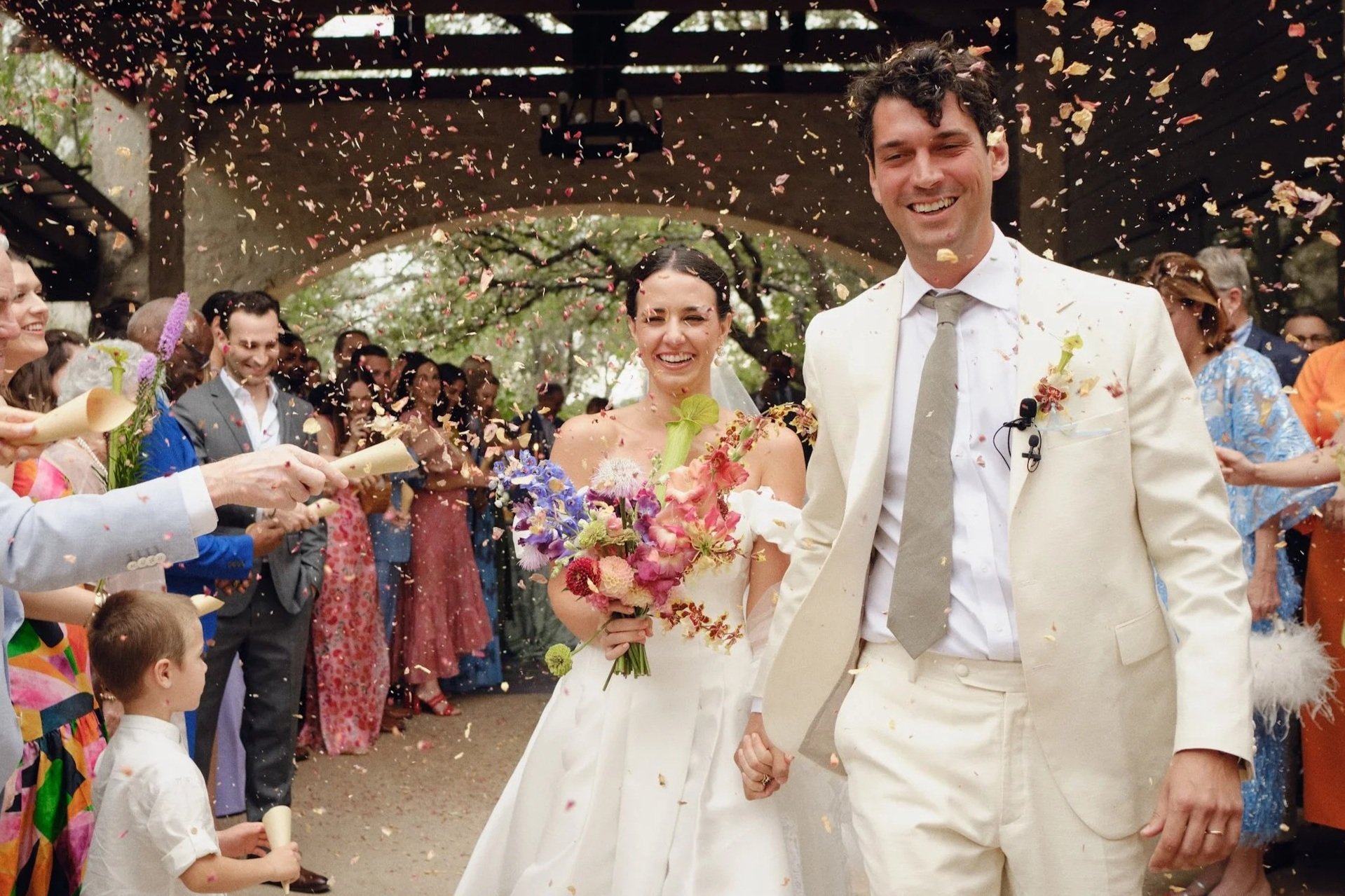 A bride and groom are celebrating their wedding as they walk hand in hand through a crowd of wedding guests who are throwing flower petals and confetti.