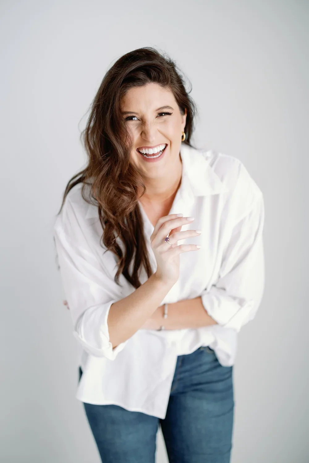 A woman with brown hair, smiling and laughing, wearing a white shirt and jeans, standing against a plain light gray background.